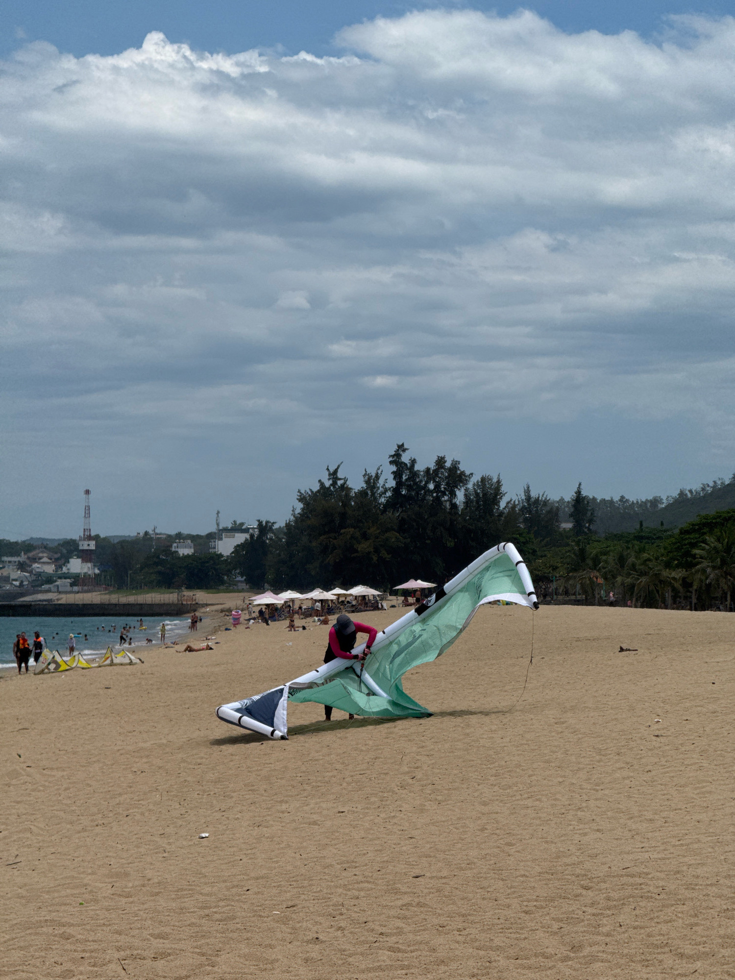 The image depicts a beach scene in Nha Trang, Vietnam. In the foreground, a person in a pink wetsuit is adjusting a large, light green and white kite on the sand. The kite is partially laid out, with its fabric spread on the beach. In the mid-ground, there are other kites on the sand, as well as several people walking and sunbathing. Several beach umbrellas are set up, providing shade. The ocean is visible in the background, with small waves and a few people swimming. A pier or breakwater separates the beach from the town. In the distance, the city of Nha Trang is visible, with buildings, trees, and a distinctive red and white radio tower. The sky is partly cloudy with a mix of blue sky and grey clouds.