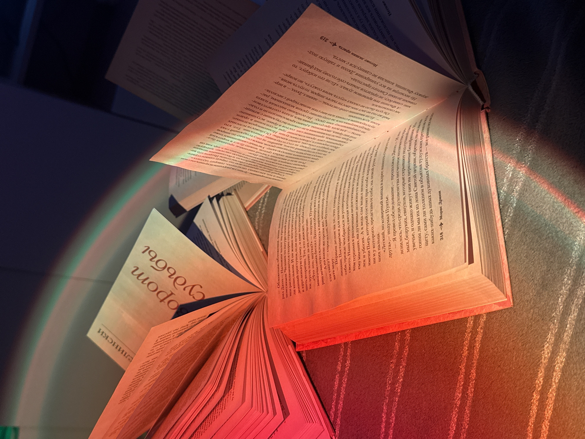 A close-up shot captures the pages of an open book, illuminated by a rainbow light effect. The book appears to be in Russian, its pages filled with text. The book's spine is red. It rests on a surface, possibly a striped blanket or carpet, adding texture to the scene.