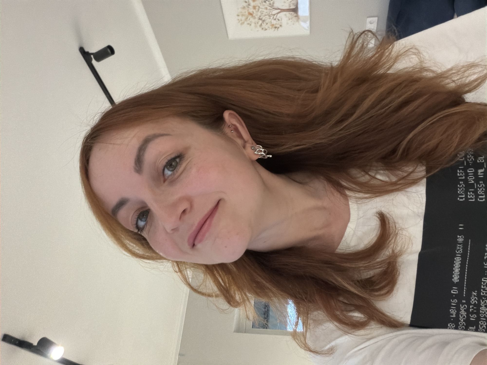 This image, captured in Chaikovsky, Russia, features a young woman with long, wavy reddish-brown hair. She is seen from a low angle, looking upwards towards the camera with a subtle, pleasant smile. Her green eyes are directed at the viewer. She is wearing a white top, which has a black stripe across the chest area displaying various lines of white text.
Notable details include multiple ear piercings on her left ear: several small studs on the upper cartilage and a decorative, intricate silver earring on the earlobe. Her hair appears somewhat tousled and flowing.
The setting is indoors, likely a room, characterized by light-colored walls and ceiling. In the background, a light fixture is visible mounted on the ceiling or upper wall, along with a framed picture depicting a tree motif. A glimpse of a window or reflective surface and a white desk or table can also be seen. The lighting is soft and ambient, suggesting indoor conditions.
The visible text on the black stripe of her shirt, possibly a design or label, includes: "11 501XX910000000-10-9118M-88", "9945045=", "DL 16 77 99%.", "958193045 FCFS111", "CLASS=LEFI COL", "LEFI VOID <SPA", and "CLASS INL BL". This text appears to be technical or coded information. The overall impression is a casual, relaxed selfie or portrait taken indoors.