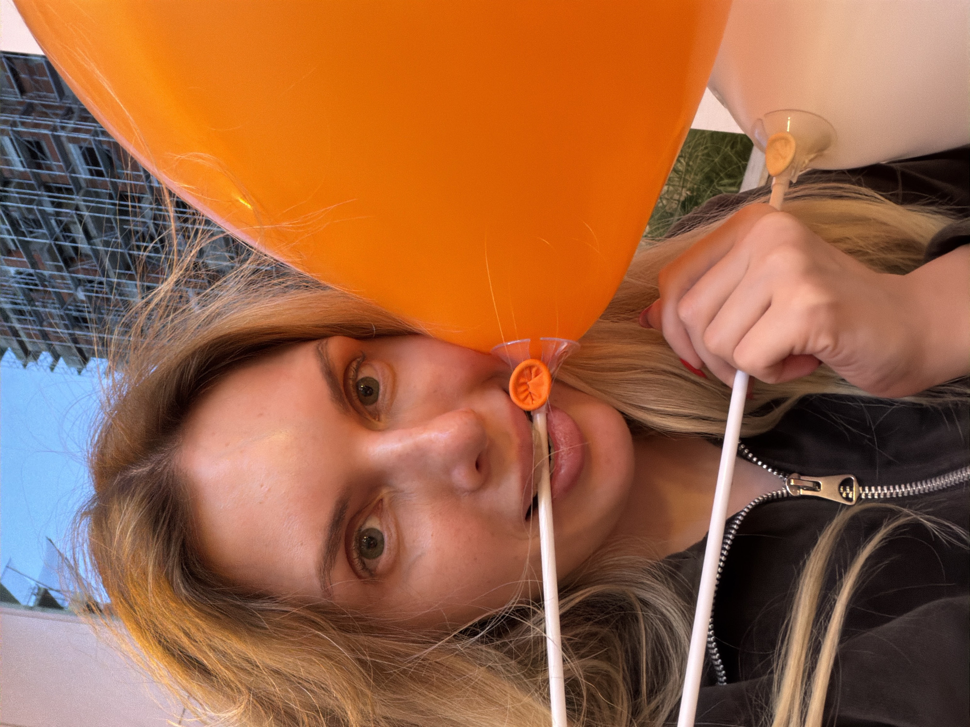 A blonde woman with visible freckles is holding an orange balloon and a white balloon close to her face. The orange balloon is positioned near her eye, with the plastic stick extending towards her mouth. She is wearing a dark-colored zip-up jacket, and the background shows a blurred cityscape, suggestive of an urban environment. The image could have been taken in the city of Budva, Montenegro, based on the potential building design visible in the background. Further forensic details may be derived from a higher resolution picture and additional angles.