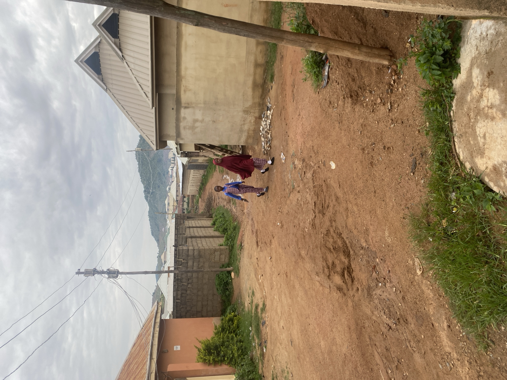In Jos, Nigeria, two children are walking down a dusty dirt road lined with simple buildings. The child on the left wears a blue and red checkered shirt and pants, while the child on the right is covered in a maroon hijab and patterned pants. Behind them, brick walls and beige-colored buildings with distinctive peaked roofs are visible under a cloudy sky. Power lines stretch across the scene, supported by a wooden pole. The road is uneven with patches of grass growing along the edges.