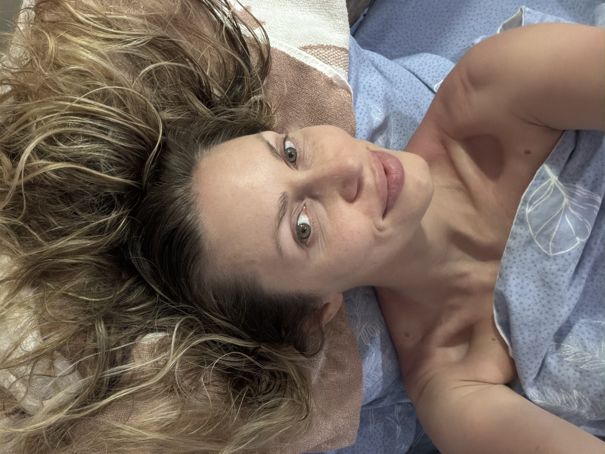 A woman with blonde hair is lying on her back, looking at the camera. She is wearing a blue patterned sheet or blanket that covers her chest and shoulders. Her hair is spread out around her head. The lighting suggests an indoor setting, possibly a bedroom. The image does not contain any information that would allow for its location to be determined in the city of Sochi, Russia.