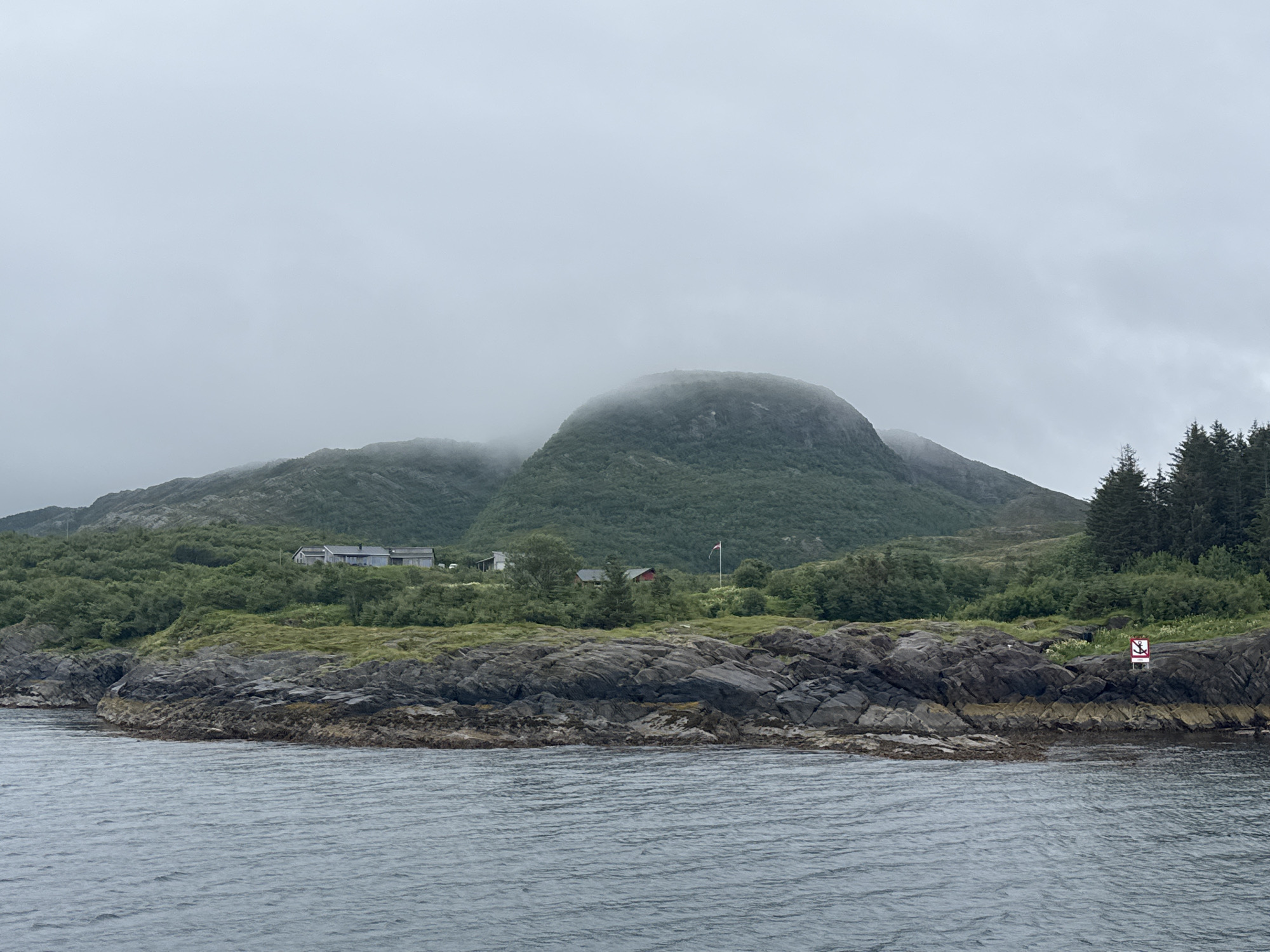 This image shows a rocky coastline with lush green vegetation leading up to a mountain shrouded in fog. Several buildings are visible nestled into the hillside, along with a Norwegian flag on a pole. The water in the foreground is choppy under an overcast sky. A red and white sign with an anchor symbol is visible on the rocks in the lower right corner of the image. The location is identified as Demmahågen, Norway.