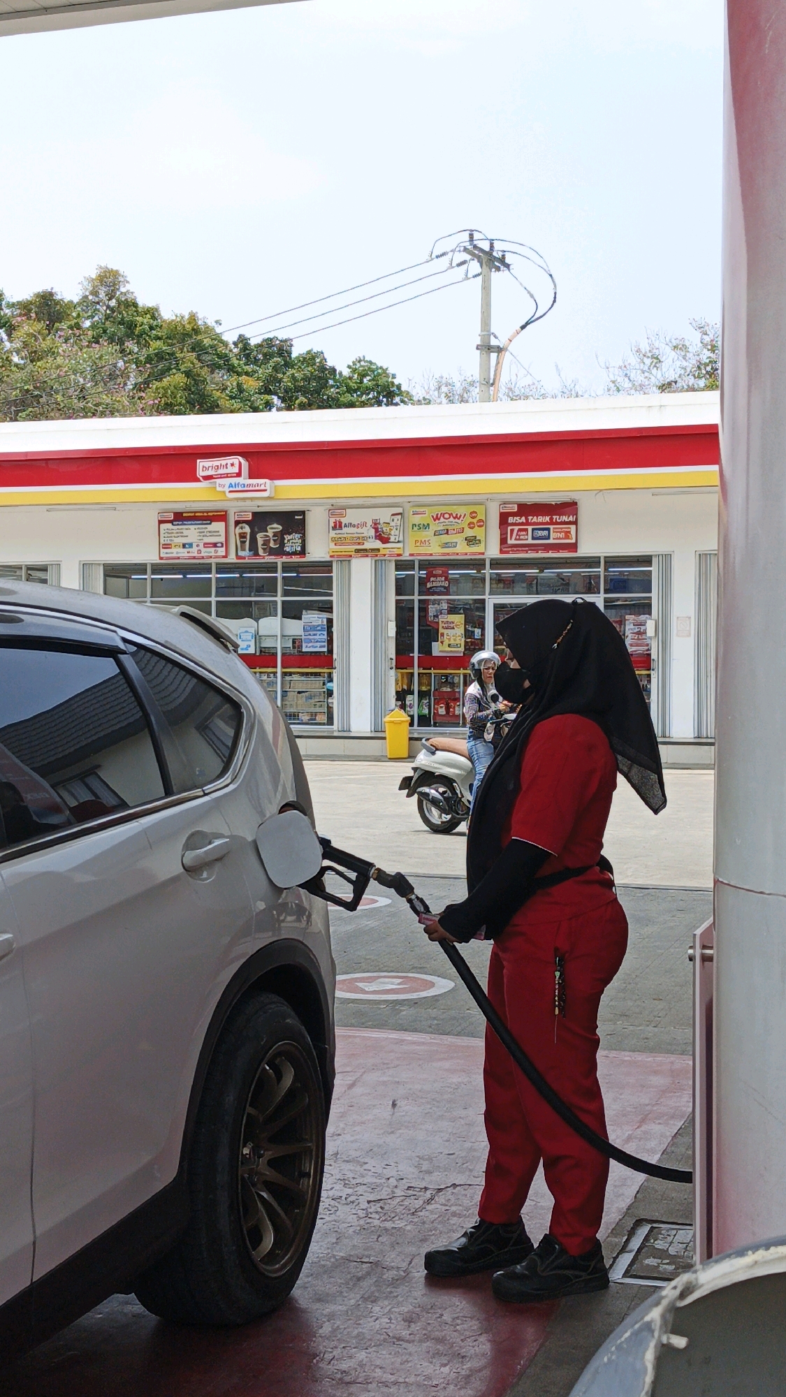 A woman wearing a red uniform and black hijab is seen from the side at a gas station, holding a fuel nozzle as she refuels a white SUV. She has a black face mask on. The gas station has a red and yellow awning, and a building with signage is visible in the background. A person on a scooter is also present in the background, as well as trees and a utility pole. The ground is paved with red and gray concrete. The scene is located at a gas station in Desa Wonoharjo, Indonesia.
