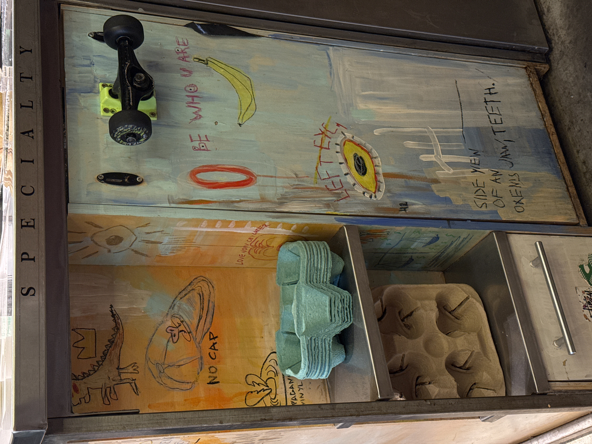 This media file presents a collection of three vertically stacked, open display compartments within a metal and wood frame. The scene features a blend of artistic decorations, everyday objects, and handwritten text, suggesting an eclectic and creative display, possibly in an art space, a shop, or a personal studio in Moscow, Russia.
The top compartment features a black skateboard truck with black wheels mounted on a neon-green base. The back wall is painted light blue and white, adorned with a yellow banana drawing, a red oval, and an abstract yellow and black eye design with radiating lines. Handwritten text includes "BE WHO YOU ARE," "42," and "SIDE VIEW OF AN OXEN'S JAW, TEETH."
The middle compartment, painted in warm orange and yellow tones, contains a stack of light green molded paper egg cartons. Its back wall is decorated with a cartoonish dinosaur wearing a crown, a drawing of a baseball cap with a face, and text that reads "NO CAP," "LOVE NOW CRY LATER," and "PAGAN VINYL." A sun-like drawing with an avocado in its center is also visible.
The bottom compartment is partially visible, showing a light brown molded paper egg carton and a silver metal drawer handle. A vertical metal panel on the far left side of the frame displays the word "SPECIALTY." No human subjects are present, and no specific activity is taking place; the image captures a static display.