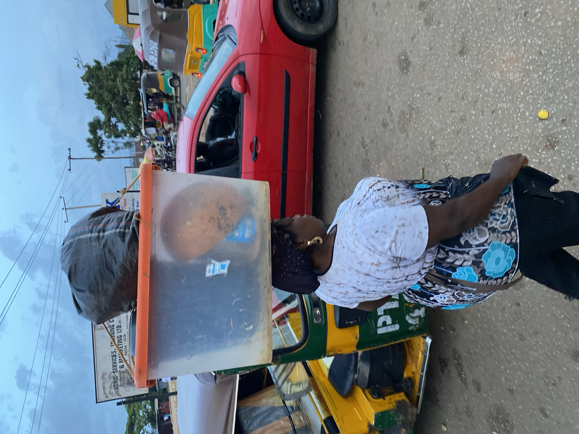 In Bukuru, Nigeria, a woman carries a large plastic container filled with items on her head. The container is balanced atop a dark plastic bag. She is walking next to a yellow and green auto-rickshaw. Behind her, a red car is parked on the street. The sky is overcast, and power lines are visible overhead. A sign in the background advertises "AGRIC SERVICES, TRAINING C & MARKETING LTD."