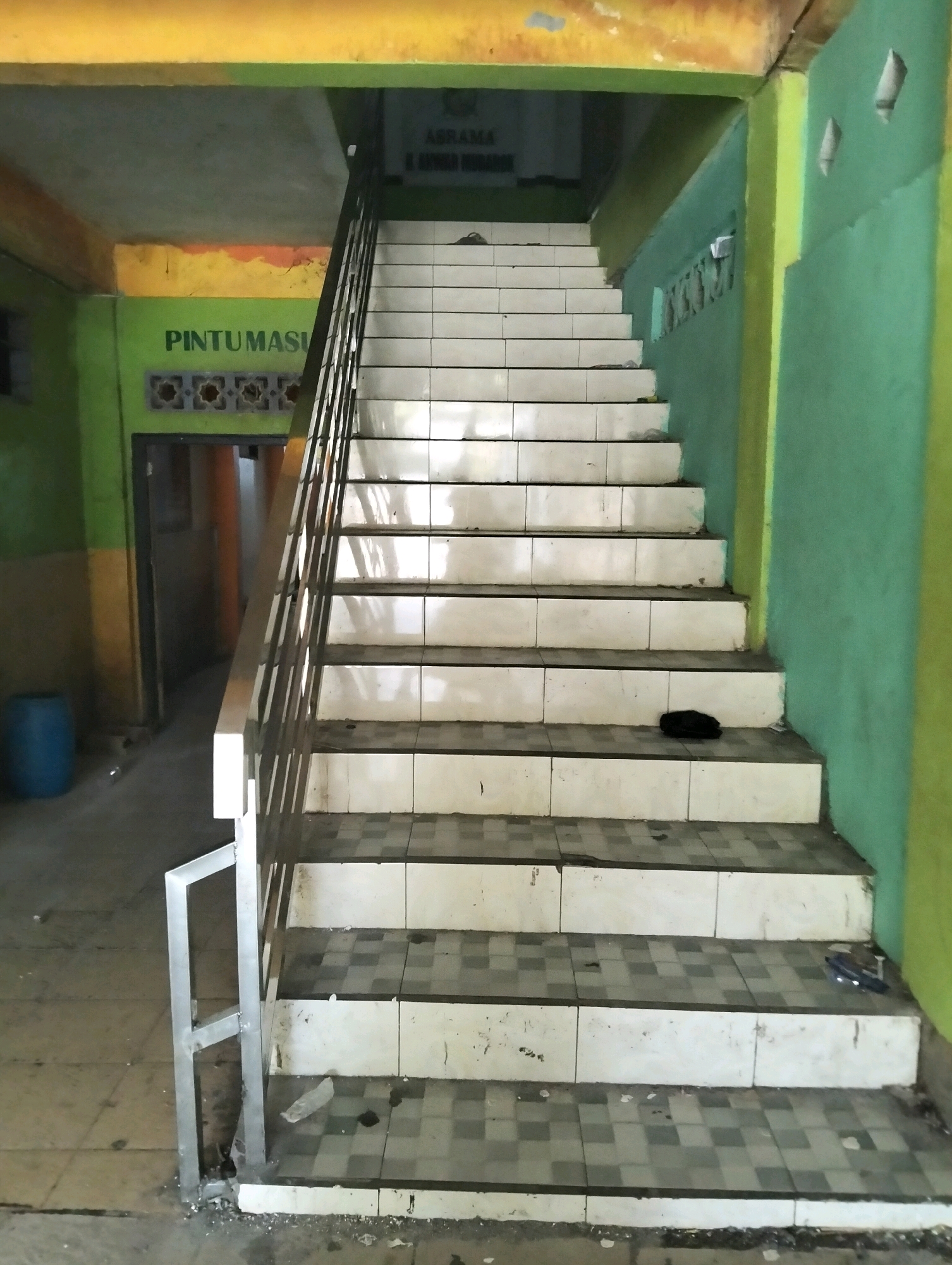 A perspective shot looks up a tiled staircase in a building with brightly painted green and yellow walls. The stairs are made of white tiles with a checkered pattern on the risers. A metal railing runs up the left side of the stairs. To the left of the stairs, a blue plastic barrel is visible. Above the entrance on the left, the word "PINTUMASI" is painted in green letters. Above the top of the stairs, text is visible on the wall, reading "ASRAMA". The lighting suggests an indoor, possibly public or institutional space.
