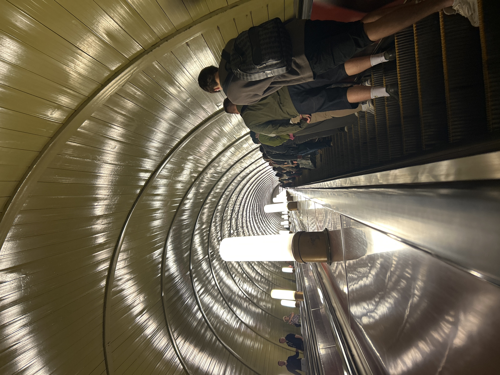 The image shows a long escalator tunnel with a reflective, metallic ceiling that curves into the distance, creating a sense of depth. Several people are riding the escalator, some standing and others with bags. The escalator steps are dark and ribbed, with yellow markings.
The tunnel is illuminated by a series of bright, cylindrical lights mounted on the wall. The metallic surfaces of the ceiling and walls are highly reflective, catching the light and creating streaks of light and shadow that accentuate the tunnel's curvature. The overall impression is one of a modern, possibly underground transportation system.
The people on the escalator are dressed casually, suggesting it could be daytime or a busy period for travel. The lighting and the architecture suggest an indoor, urban environment. The image is taken from a low angle, looking up the escalator, which emphasizes its length and the vastness of the tunnel.