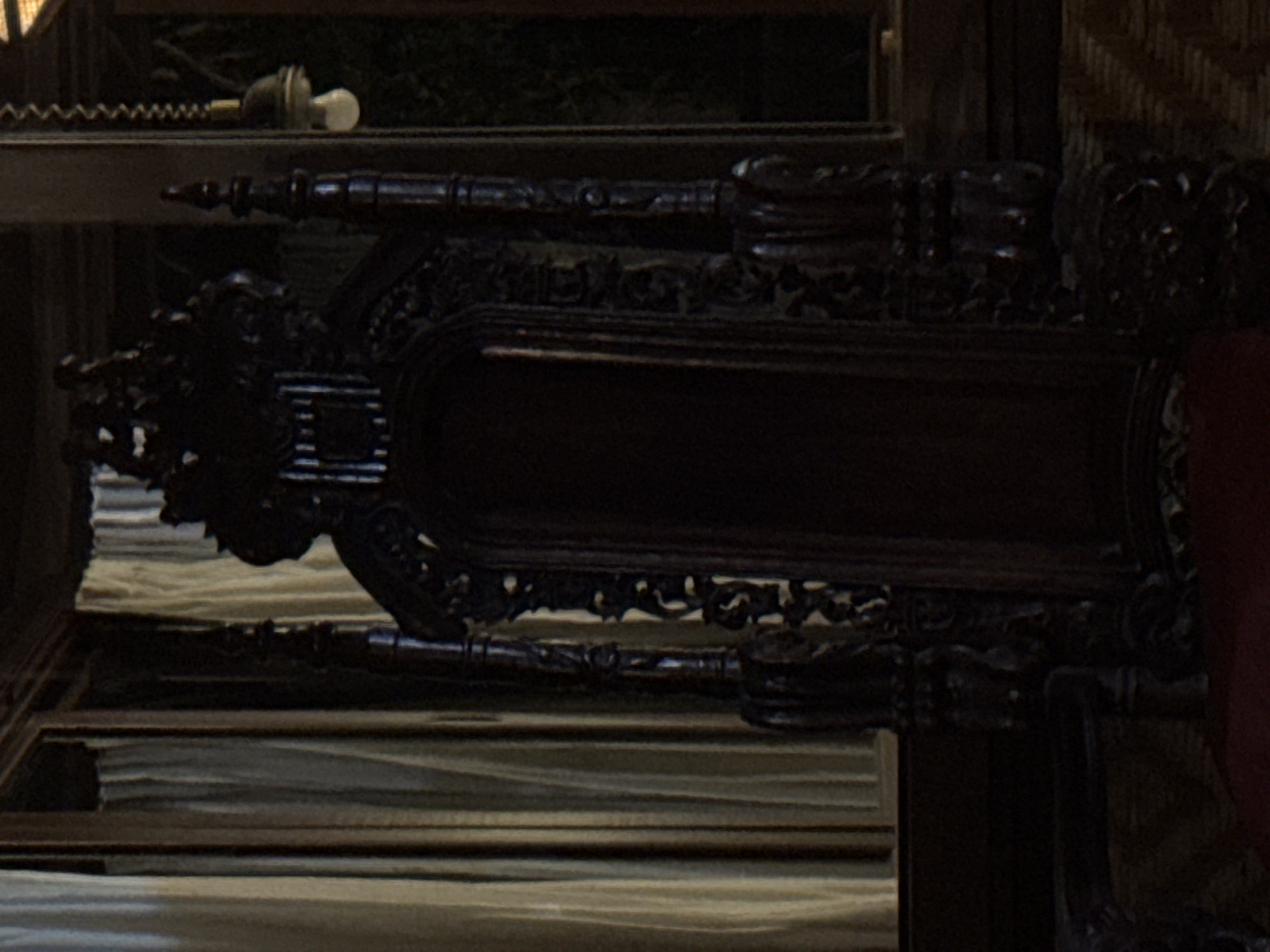 The image shows a close-up of an ornate, dark wood structure in what appears to be an interior setting. The woodwork features elaborate carvings. A lamp or bulb is visible in the upper left corner. The location is specified as Ubud District, Indonesia.