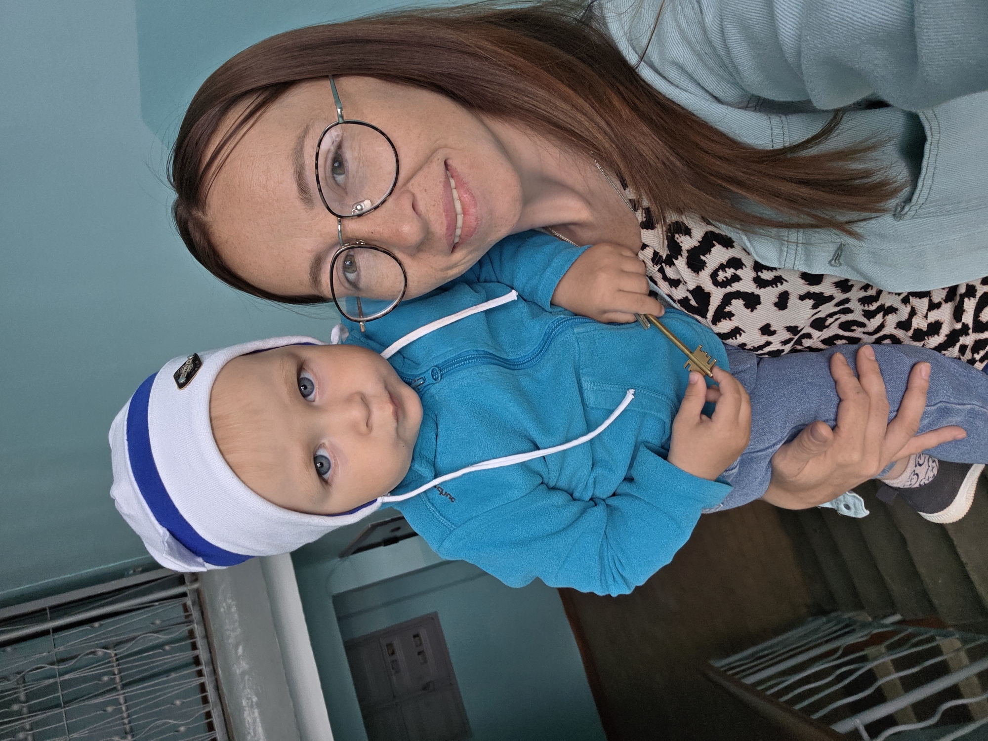 A young child with blue eyes and fair hair, wearing a blue hooded jacket and a white and blue striped hat, is held by a woman with long brown hair and glasses. The woman is wearing a patterned top and a light blue denim jacket. They are standing on a landing in what appears to be an apartment building stairwell, with a metal railing and concrete stairs visible. The walls are painted a light blue. The child is holding a small brass-colored key.
