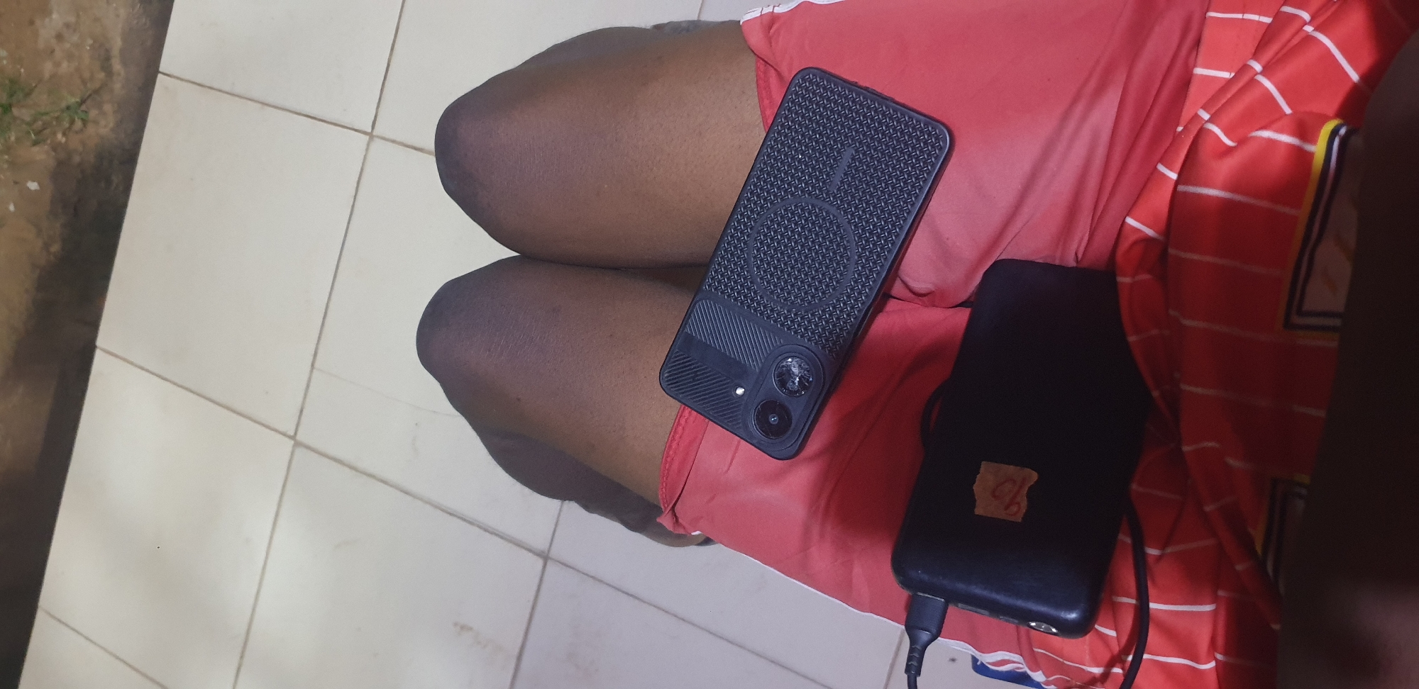 The image depicts an overhead view of a person seated in Narayi, Nigeria, with their lower body from the thighs to the knees visible. The individual has dark-toned skin and is wearing red shorts with white vertical stripes and a partial logo or design on the lower left. Resting on their lap are a black smartphone and a black power bank. The smartphone is enclosed in a textured black case featuring a prominent circular pattern and a patterned grip. Its dual camera module is visible, with one lens showing apparent damage. A black cable connects the smartphone to the power bank, indicating active charging. The power bank has a small, light-brown adhesive label affixed to its top surface. The immediate environment consists of a clean white tiled floor with grey grout lines. Beyond the tiled area, an unpaved outdoor surface with dark soil and sparse green vegetation is visible.
