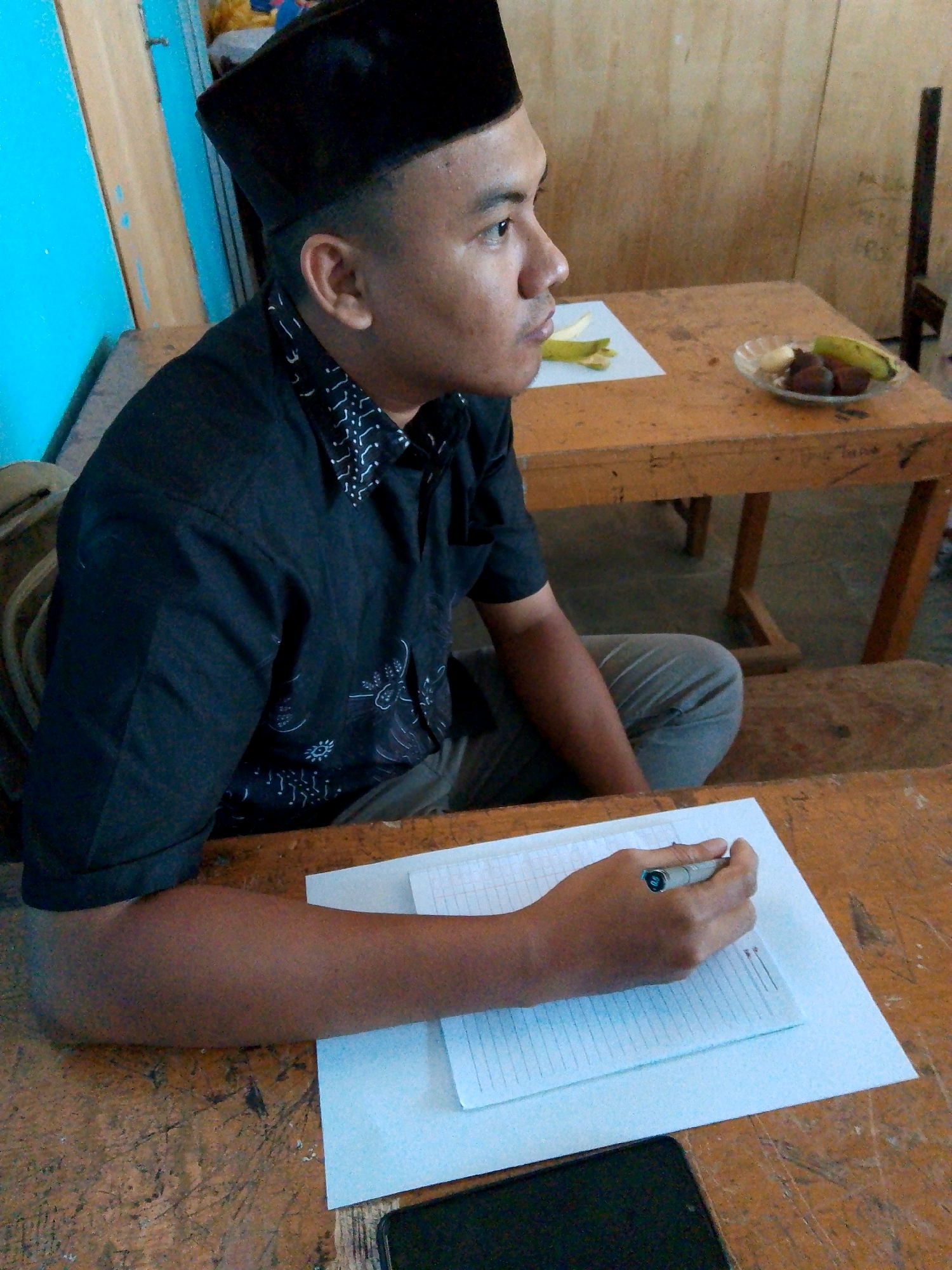 A medium shot captures a young man wearing a black peci hat and a black patterned shirt, seated at a wooden desk. He is looking to his right with his head angled slightly downwards. His right hand, holding a pen, is positioned over a notepad on the desk in front of him. To the right of the notepad, a smartphone is visible. The background reveals another wooden desk with a plate of fruit and banana peels. The environment suggests an indoor setting, possibly a classroom or a communal space, with visible wooden structures and a blue-painted wall on the left. The overall lighting is natural, and the scene appears to be set during the daytime. The context of Kuripan Utara, Indonesia, is noted but not visually evident in the image itself.