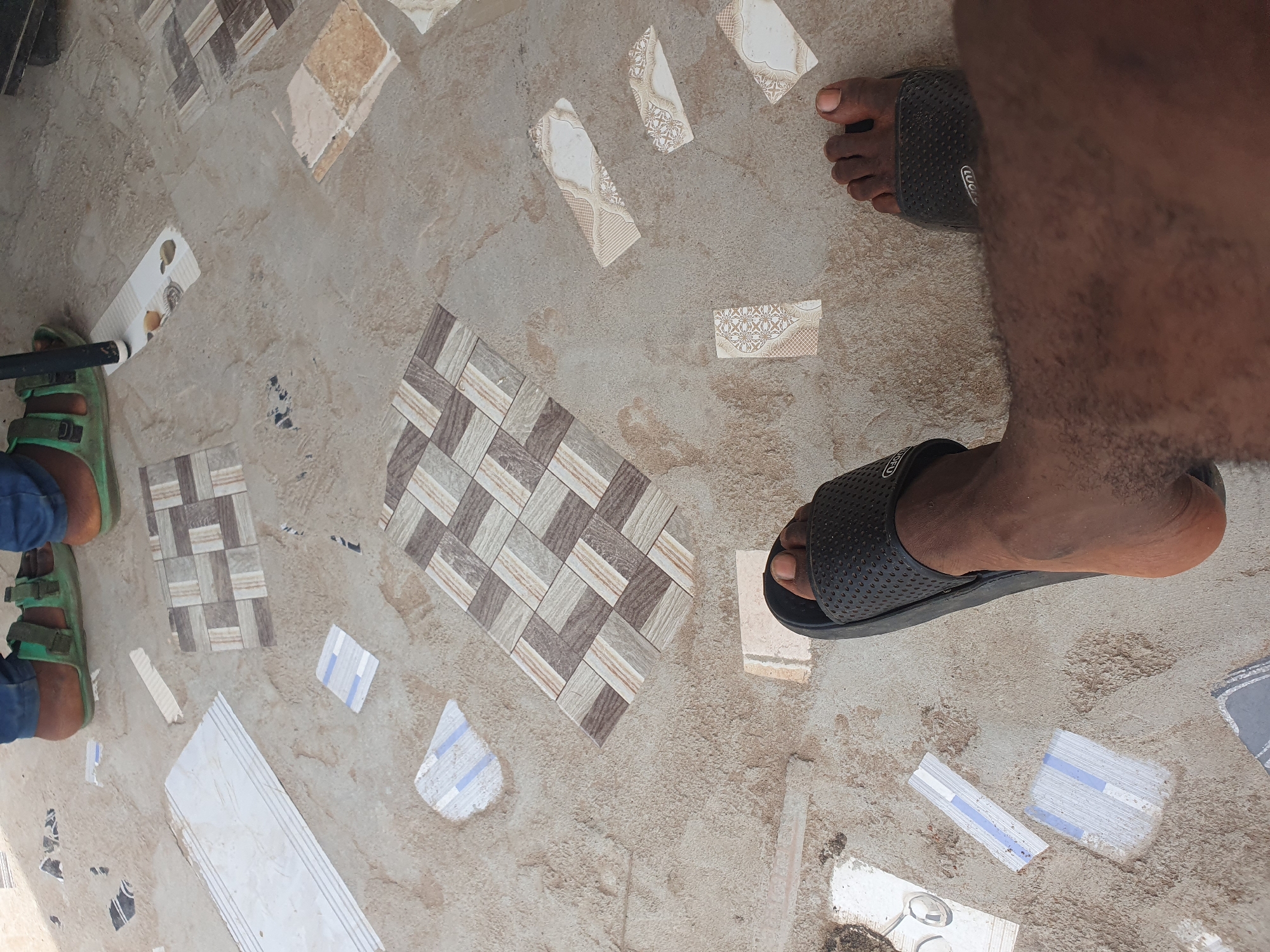 This media file captures a close-up, overhead view of a person's legs and feet standing on a concrete floor with scattered tiles. The setting appears to be an outdoor or unfinished area, possibly a construction site or a poorly maintained patio, given the scattered tiles and rough concrete. The person is wearing green sandals on their left foot and black slip-on sandals on their right. The tiles are varied, with some having geometric patterns in shades of brown, beige, and grey, while others are plain white with decorative elements or blue stripes. Some tiles appear to be broken or misaligned, suggesting ongoing work or damage. The time of day is not definitively indicated, but the lighting suggests it is daytime. There are no clear indicators of weather or emotions. The scene is likely in Lokoja, Nigeria, as per the provided location context. There is some faint text visible on the black sandal strap.