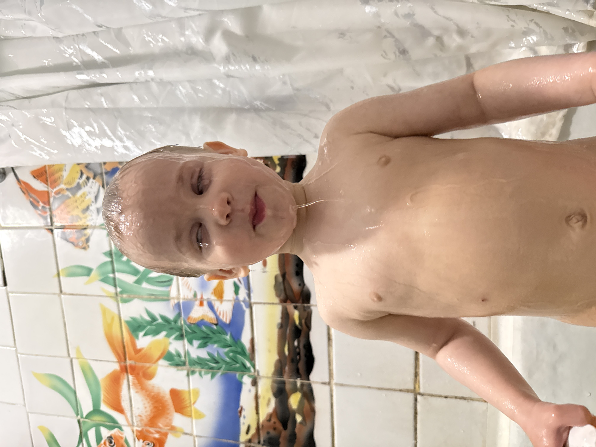 A close-up, rotated portrait captures a young child during a bath or shower. The child, likely a toddler or preschooler, is seen from the chest up, with water dripping over their fair skin and light hair. Their eyes are mostly closed, and a faint smile plays on their lips, suggesting enjoyment or a reaction to the water.
The setting appears to be a bathroom. In the background, a white, translucent shower curtain is visible to one side, showing crinkles and folds. To the other side, a tiled wall provides a colorful backdrop. The tiles feature various decorative motifs, including orange goldfish surrounded by green foliage, butterflies, and what appear to be white birds (possibly cranes) against a blue and dark background. Some grout lines between the tiles are visible. The child's body is covered in water droplets, highlighting the activity.
The image conveys a moment of innocence and playfulness during a hygiene routine. Given the provided context, the scene takes place in Rusanovo, Russia. There is no visible text in the image.
