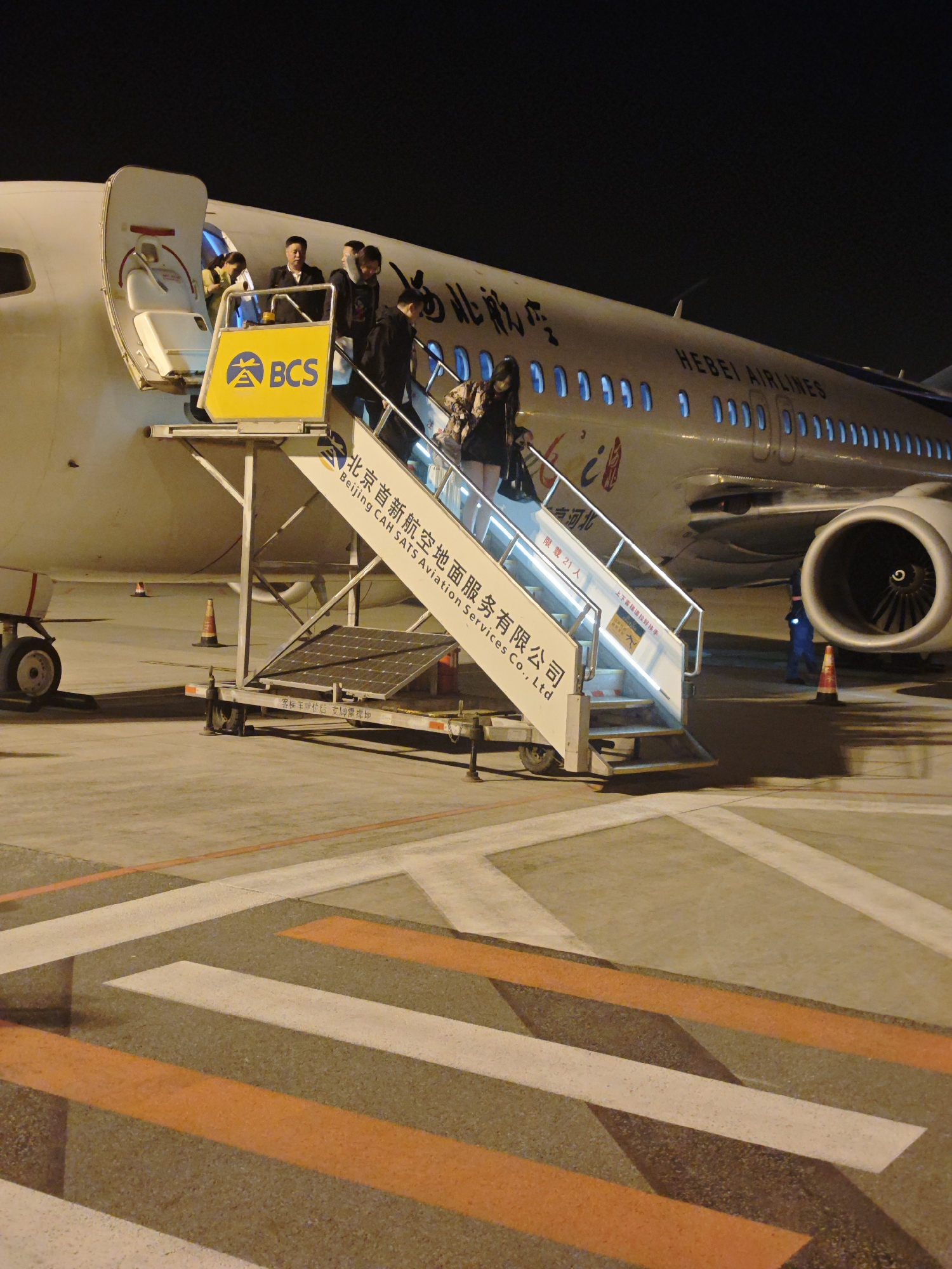 The image depicts an nighttime scene at an airport tarmac in Langfang, China, where passengers are disembarking or boarding a "HEBEI AIRLINES" aircraft. The white jet, featuring Chinese characters "台河北" on its fuselage, is partially visible, with one engine on the right side.
A mobile air stairs unit is positioned against the open aircraft door. Several people, including what appears to be a flight attendant, are visible on the stairs and in the doorway. The air stairs prominently display a yellow "BCS" logo and branding for "北京首新航空地面服务有限公司" (Beijing CAH SATS Aviation Services Co., Ltd). Textual instructions in Chinese are visible on the stairs, including "上下客梯请拉好扶手" (Please hold the handrail when going up and down the passenger stairs), "限载21人" (Limited to 21 people), and "客标车位后支牌需撑地" (The rear support plate of the passenger stairs vehicle needs to be propped on the ground). Solar panels are mounted on the base of the stairs.
The ground is marked with white and orange lines, indicative of airport operational areas. A ground crew member in a reflective vest is visible near the aircraft's engine. The setting is clearly after dark, with artificial lights illuminating the scene, highlighting the activity of passenger movement between the plane and the ground.