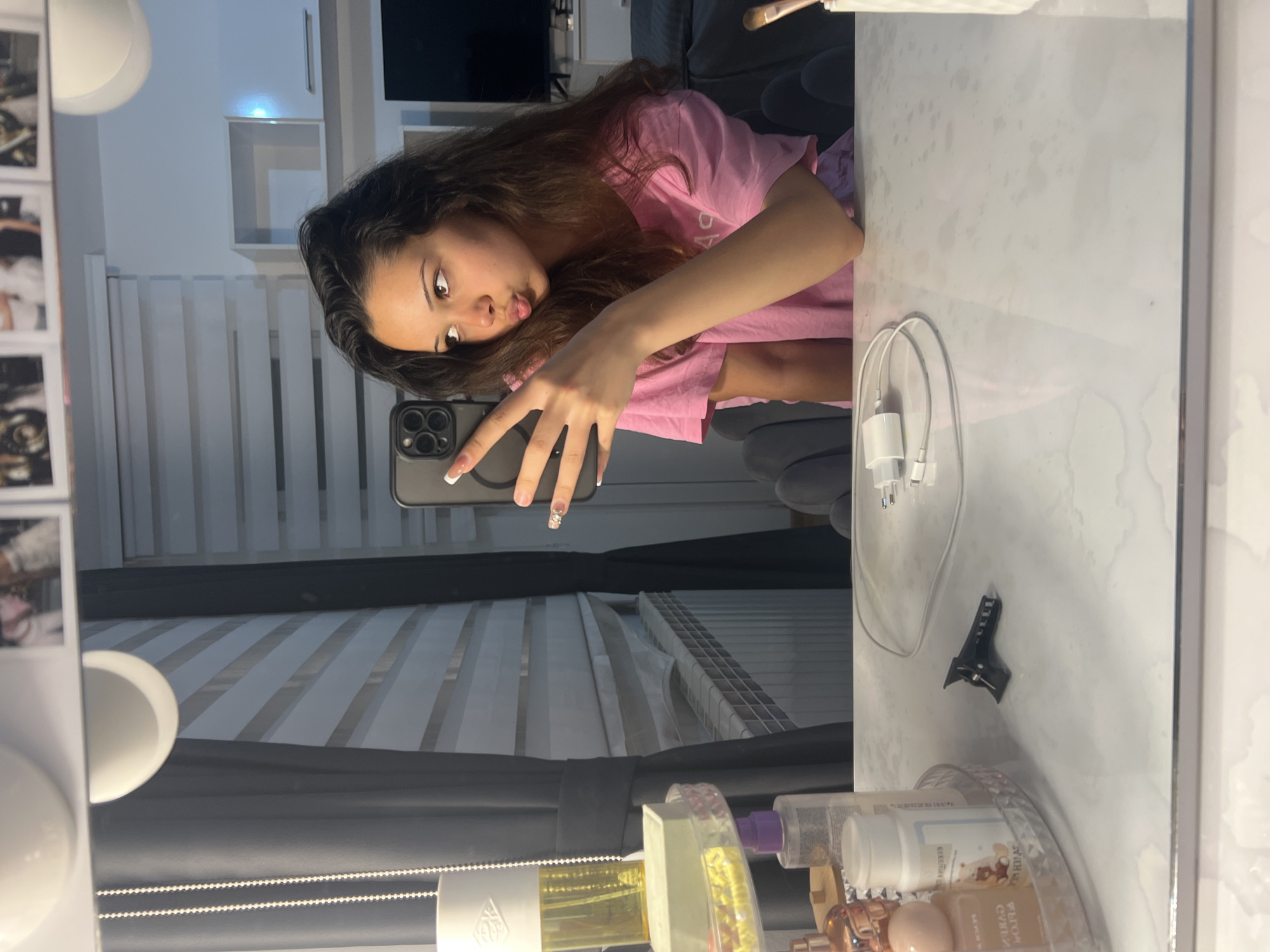 A young woman takes a mirror selfie in what appears to be a bedroom or dressing area. She is positioned on the left side of the reflection, with her upper body and face visible. She has long, dark, wavy hair and is wearing a light pink t-shirt. Her lips are slightly pursed, and she's making a "peace" or "V" sign with her left hand, which holds a black smartphone with multiple camera lenses (likely an iPhone). Her fingernails are manicured with white tips and some glittery designs.
The mirror reflects a room that includes horizontal window blinds, a dark sofa or armchair, and white wall shelves. On the frame of the mirror, to the left, several small, printed photographs are attached. Along the top and bottom edges of the mirror, circular white light fixtures are visible.
The surface in front of the mirror, likely a vanity or dressing table, is white with a subtle marble pattern. On this surface, a white charging cable with a wall adapter is coiled. To the right, a black hair clip is placed. Further to the right and foreground, a clear circular organizer holds several cosmetic or perfume bottles, some with visible labels. "PILLOW CURE" can be read on one bottle. The overall setting suggests a well-lit indoor environment, possibly during the day, with a casual and personal atmosphere.