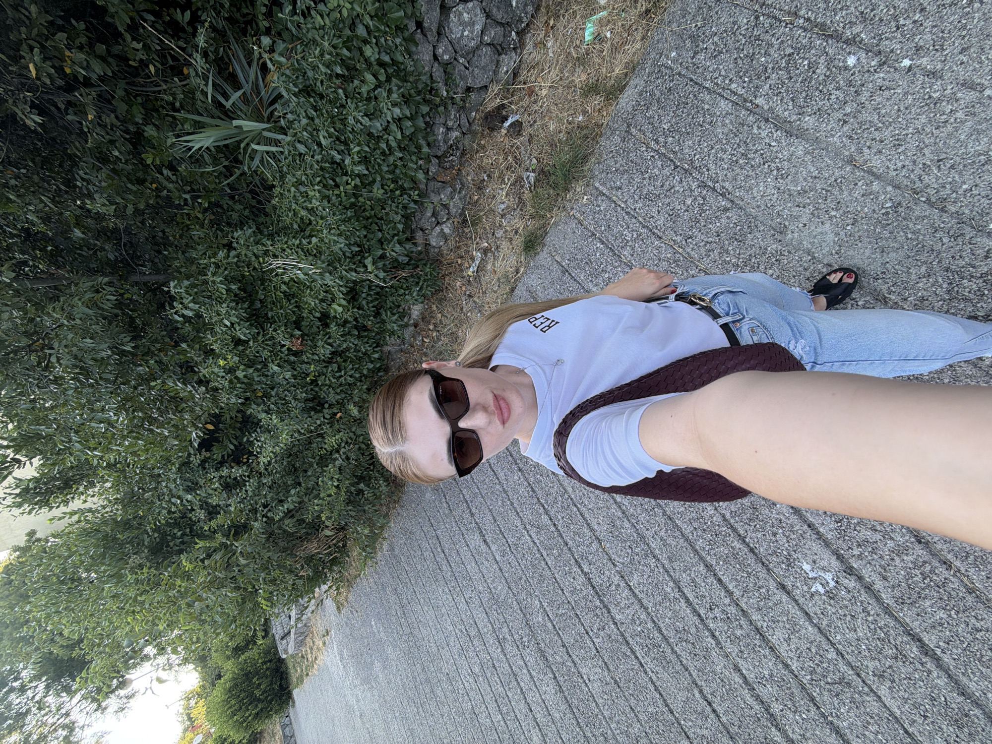 A young woman with blonde hair pulled back stands on a paved pathway next to a dense green hedge. She is wearing a white t-shirt with the letters "REP" printed on it, light blue jeans, black sandals, and dark sunglasses. She is holding a dark brown woven purse on her left shoulder and is taking a selfie with her right arm extended. The pathway is made of gray concrete with textured lines running diagonally across it. The hedge behind her is lush and green, with some dry grass and scattered debris at its base, bordering a stone wall. The scene appears to be outdoors during the daytime.
