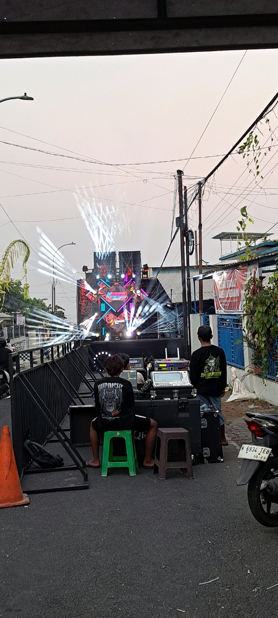 The image captures an outdoor event in Durensewu, Indonesia, featuring a large sound system setup on a stage with elaborate lighting effects. The lighting beams shoot upwards and outwards, creating a dynamic visual display against an overcast sky. In the foreground, two young men are seated behind a barrier, seemingly managing or observing the sound equipment. One man, wearing a black t-shirt with a wolf graphic and shorts, sits on a green stool. The other man, wearing a black long-sleeved shirt with a graphic and a cap, stands behind him. Several pieces of audio equipment, including mixers and speakers, are visible. A motorcycle with a license plate reading "N 6934 TE0" is parked on the right side of the frame. An orange traffic cone is positioned to the left of the barrier. The surrounding environment includes residential buildings and overhead electrical wires, typical of a street scene in the area.
