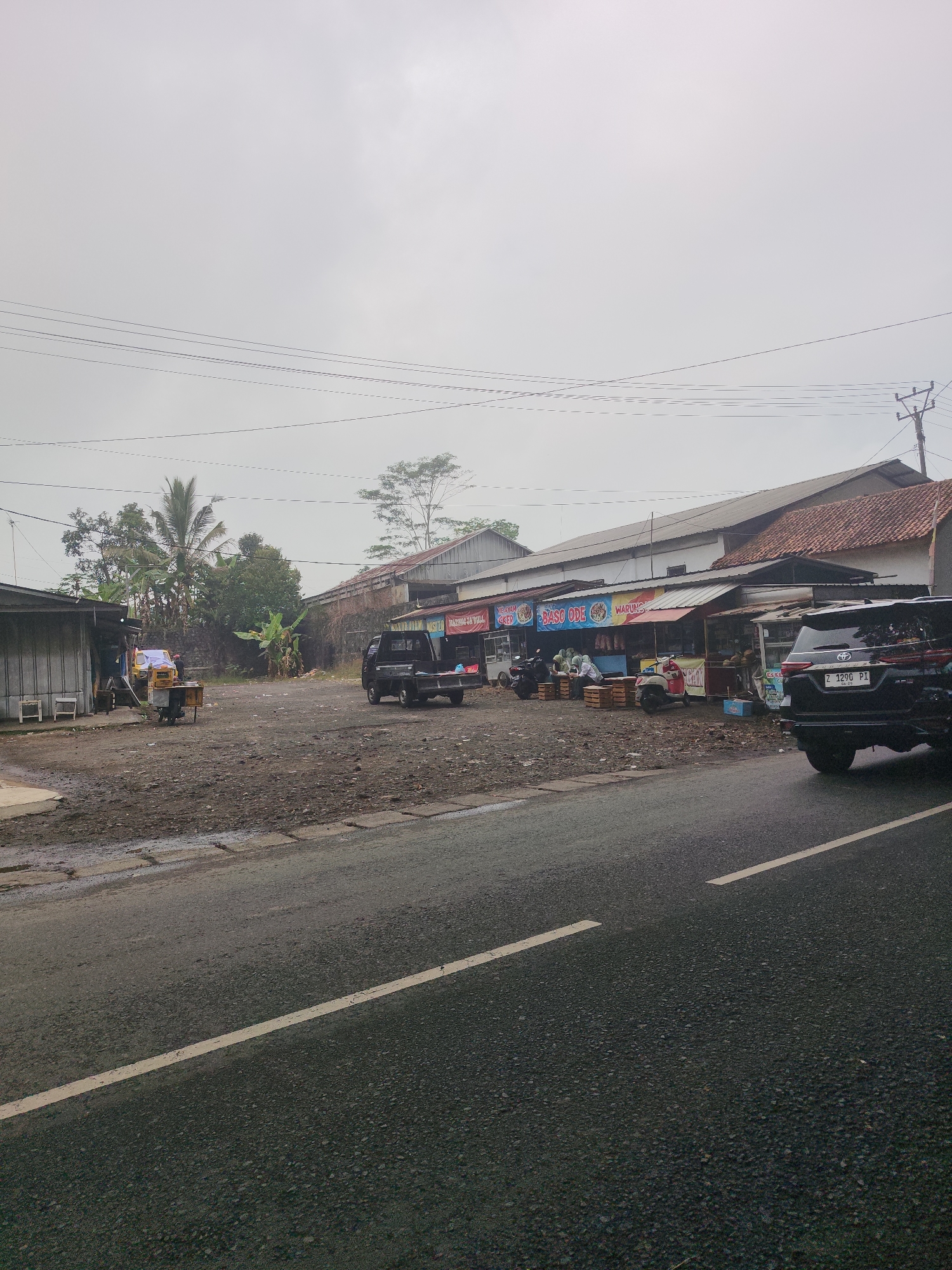 The image shows a street scene in Tasikmalaya, Indonesia, on a cloudy day. A paved road with a yellow line runs through the foreground. On the right side of the road, a black SUV is parked. Beyond the road, there are several small shops or stalls with awnings. One stall displays a sign that reads "BASO ODE". There are various vehicles parked, including a black pickup truck, a yellow mobile cart, and a few motorbikes. People are visible near the stalls, some appearing to be customers and vendors. The background features a long building with a red-tiled roof and a metal roof, along with trees and utility poles with overhead wires. The ground in front of the stalls is unpaved and appears to be dirt and gravel.