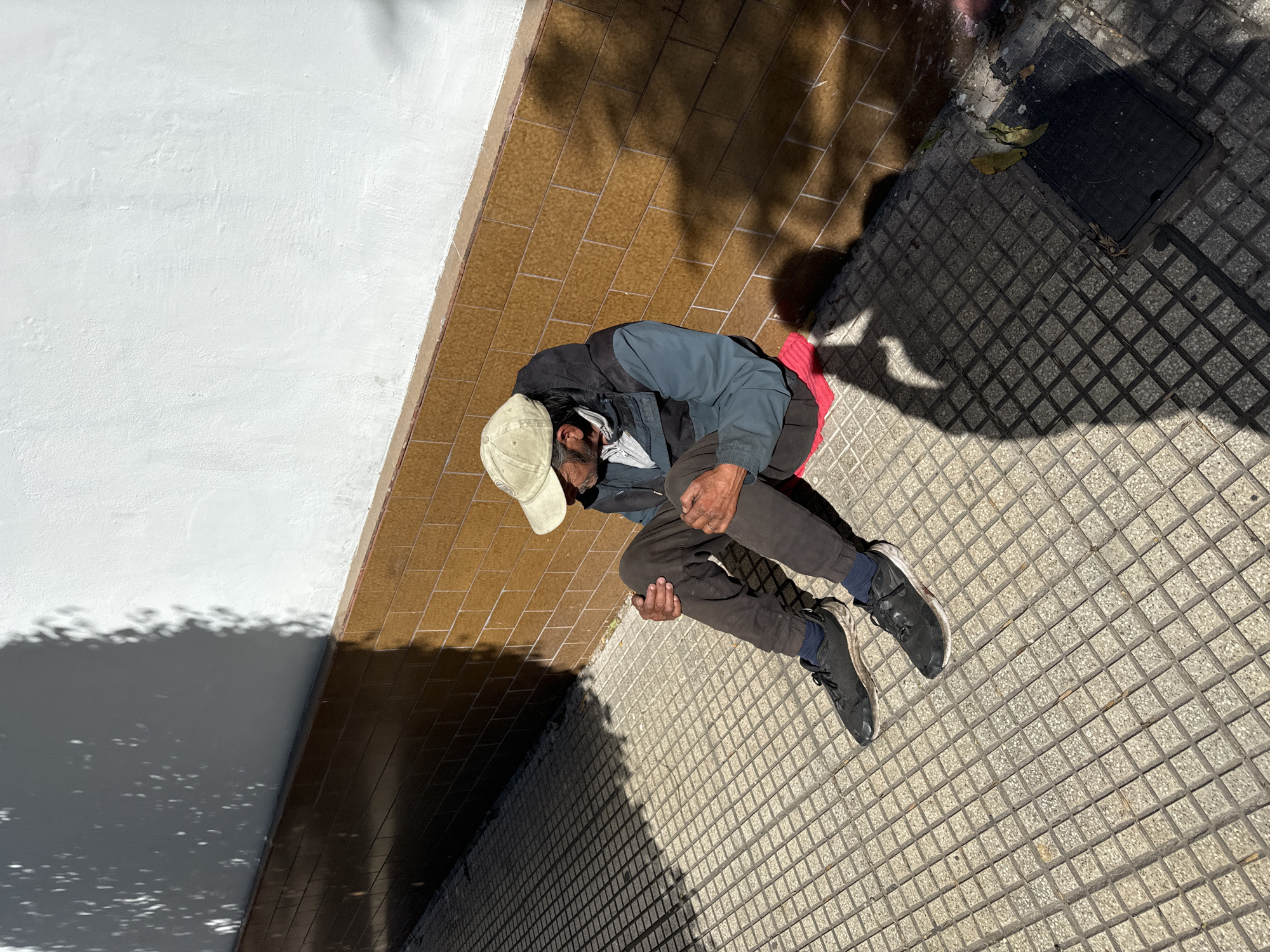 The image captures a man seated on what appears to be a red plastic crate at the corner of a building along a sidewalk. He is wearing a baseball cap, jacket, and dark trousers with black shoes. The location appears to be in Buenos Aires, Argentina, suggested by the style of the tile used. The image appears to be taken from above. The area around the seated man is paved with patterned tiles. There is a shadow of trees and other objects to the left side of the photo. A drain is nearby.