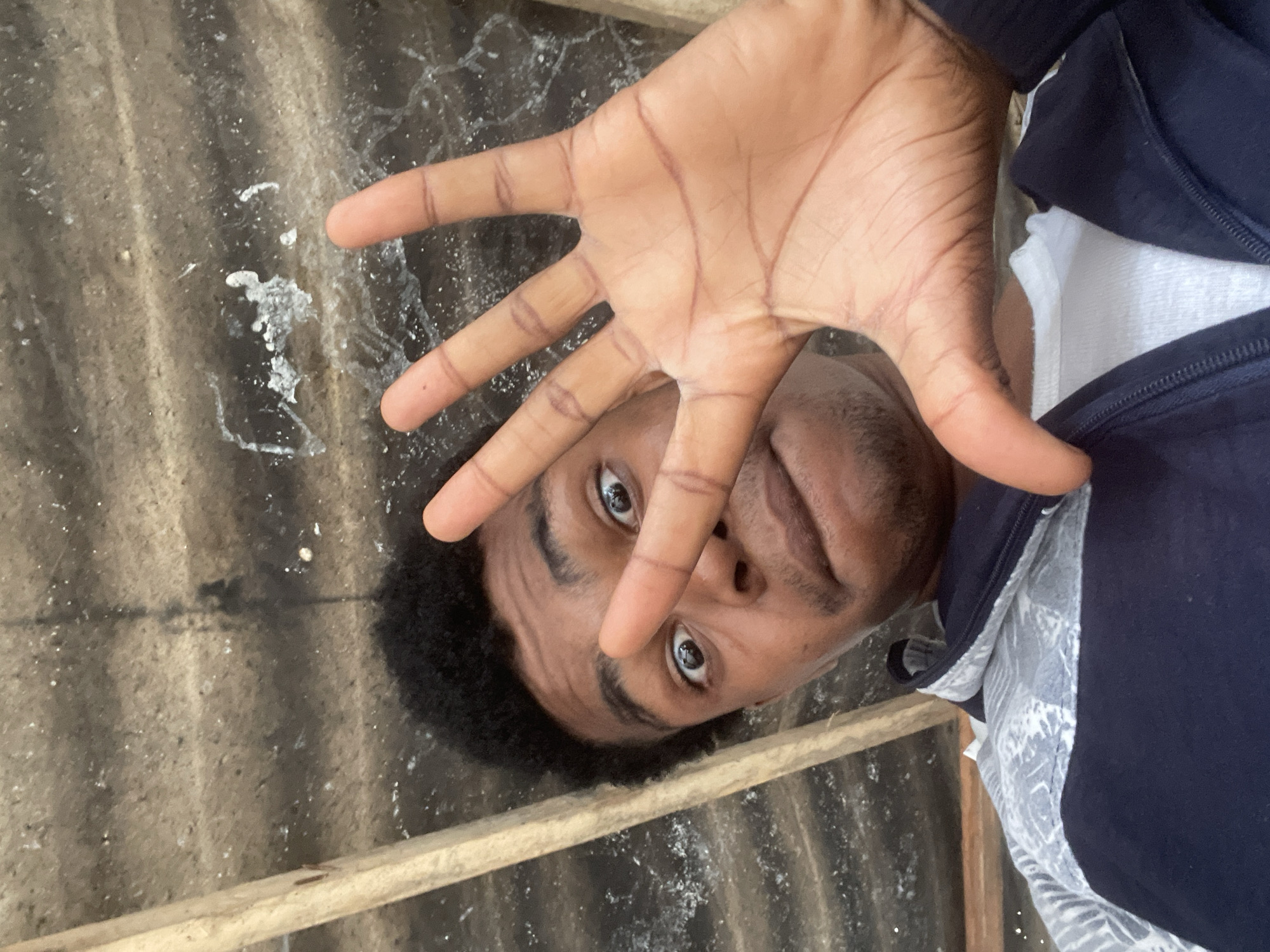 A young Black male, with dark, curly hair and brown eyes, is looking directly at the camera, with his left hand raised in front of his face, fingers splayed, blocking the view of his nose and mouth. He is wearing a dark blue zippered jacket over a white t-shirt. The background appears to be a corrugated metal roof, with wooden beams visible. The image is a selfie, taken from a low angle. The setting is likely a rural or informal urban area, possibly a workshop or storage space, given the visible construction materials and the overhead structure. The lighting suggests it is daytime.
