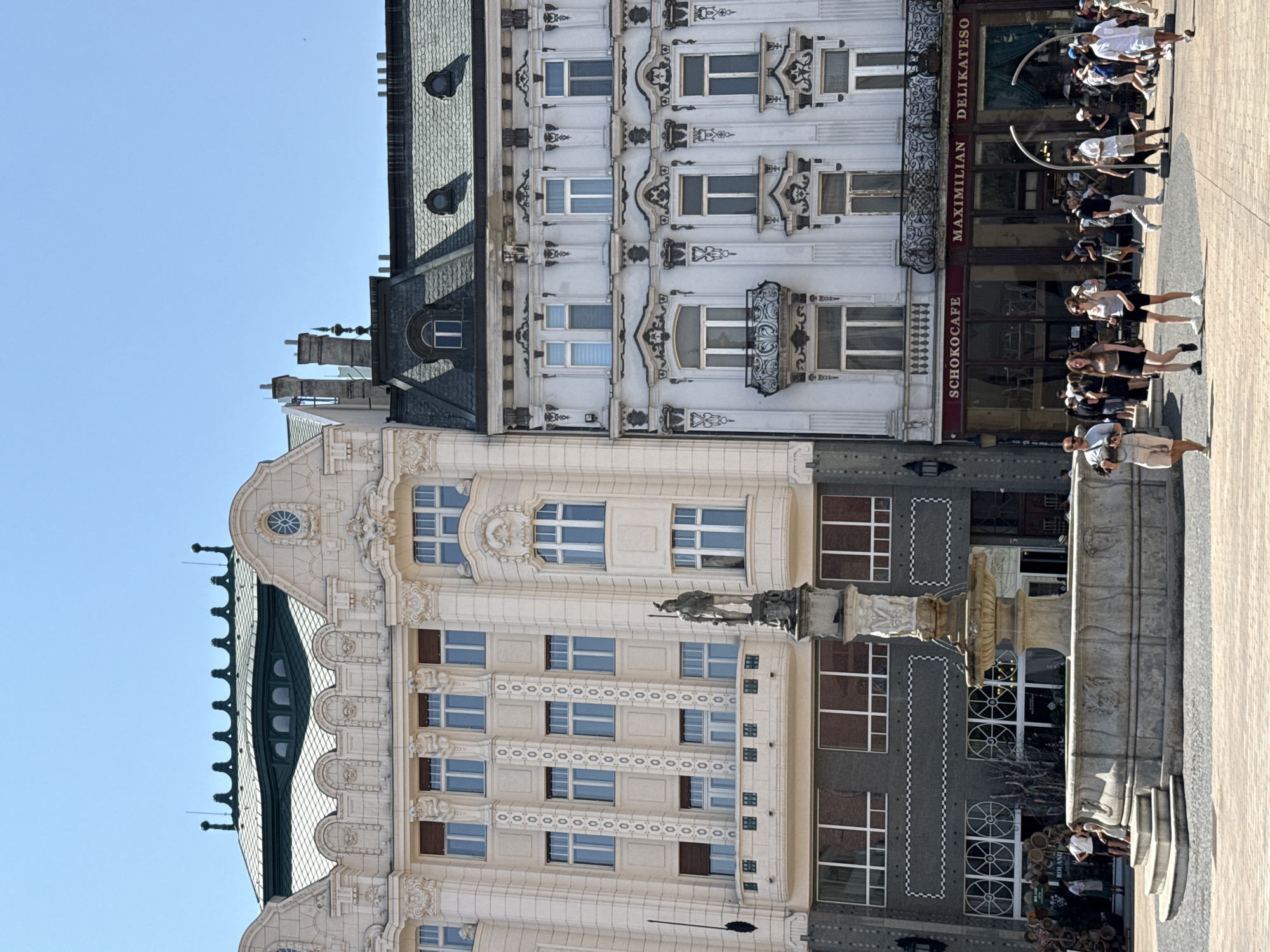 The image captures a street view in the city of Bratislava, Slovakia, likely on a sunny day, featuring prominent architecture and pedestrian activity. The foreground showcases a stone fountain with tiered basins and decorative carvings, set on a paved plaza. Several people are visible walking around the fountain and on the plaza, dressed in casual summer attire.
Behind the fountain, two ornate buildings with distinct architectural styles dominate the scene. The building on the left is lighter in color, possibly cream or pale yellow, and features intricate facade detailing, including sculpted figures, decorative friezes, and numerous windows. It has a green, multi-layered roof with arched dormer windows and decorative elements along the rooftop.
The building on the right is a lighter shade, possibly white or pale gray, with a contrasting dark green roof. Its facade is adorned with elaborate black decorative elements, scrollwork, and a prominent balcony with an ornate metal railing. This building also has many windows and appears to house a cafe or restaurant, indicated by signage that reads "SCHOKOCAFE" and "MAXIMILIAN DELIKATESO".
The sky above is clear blue, suggesting fair weather conditions. The overall scene depicts a lively urban square with historical buildings and people enjoying the public space.
