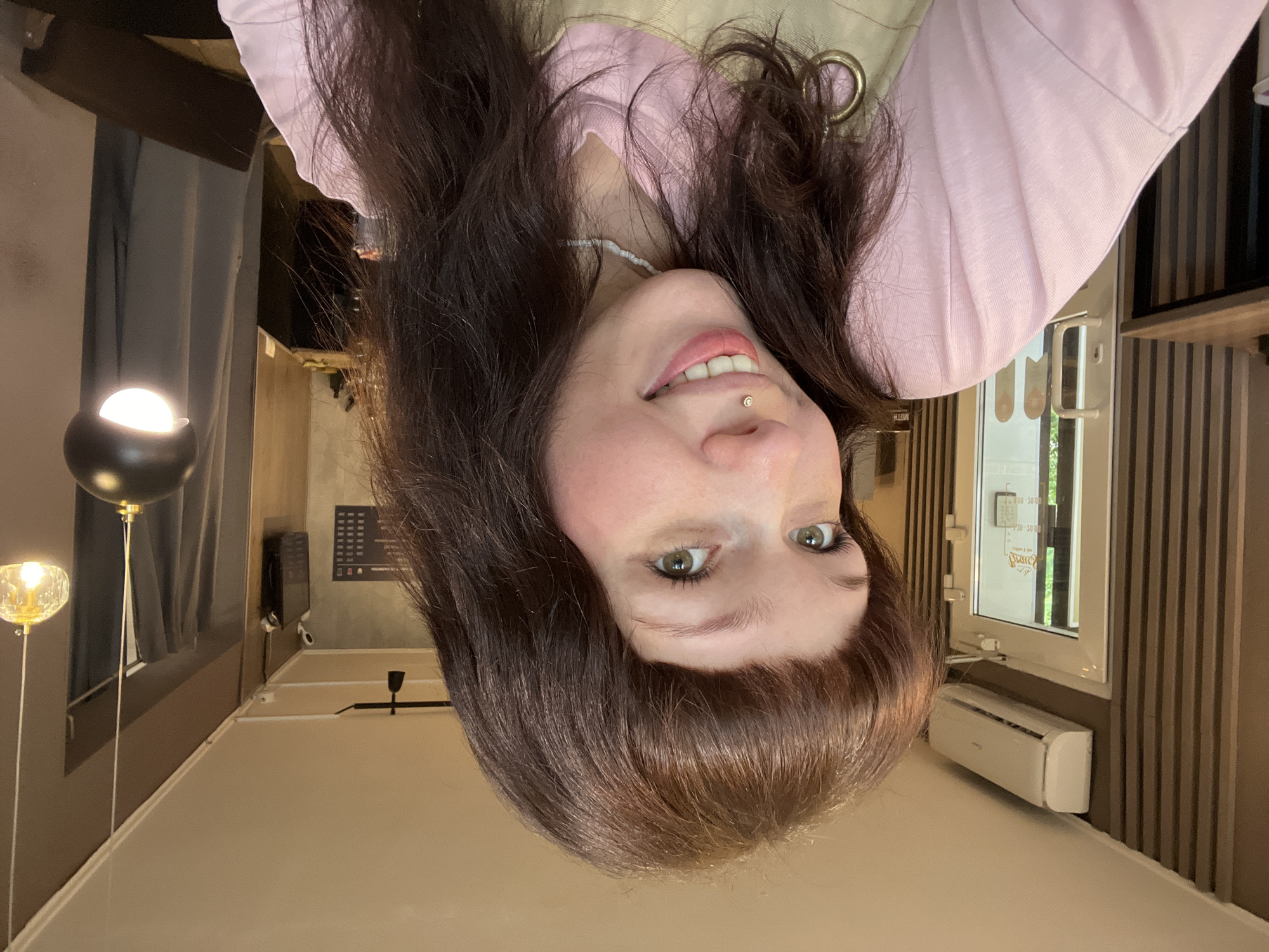 The image presents an upward-facing portrait of a young woman with long, dark brown hair, bangs, and striking green eyes. She is smiling gently and has a small, shiny philtrum piercing above her upper lip. A white beaded necklace is visible around her neck, and she wears a light pink long-sleeved top.
The setting appears to be a modern indoor space, possibly a cafe or lounge, located in Chaikovsky, Russia. The background features varied architectural elements, including dark curtains partially covering a window on the left, beside which stands a stylish black lamp with a glowing spherical light and a golden-hued decorative lamp. On the right, the wall is adorned with vertical dark wooden paneling, bordering a white-framed window that allows natural daylight into the room. Below this window is a white, wall-mounted air conditioning unit. Further back, a dark, framed sign with illegible light-colored text is visible on the wall, possibly a menu or information display. The overall ambiance suggests a bright daytime setting. The woman's relaxed posture and pleasant expression convey a calm and happy mood.