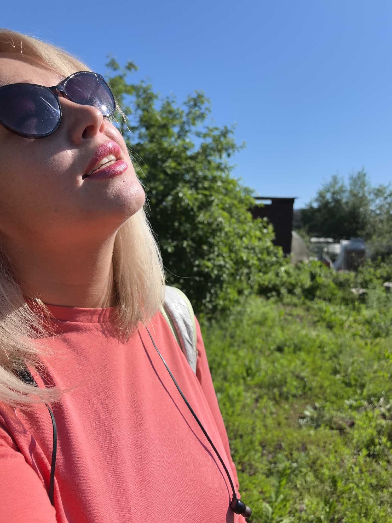A blonde woman wearing sunglasses and a coral t-shirt is looking up at the sky. She has pink lipstick on. In the background, there is a rural landscape with green vegetation, trees, and a small building. The sky is clear and blue. The scene appears to be outdoors on a sunny day, possibly in Noviy, Russia, given the presence of what looks like a dacha or small rural house.