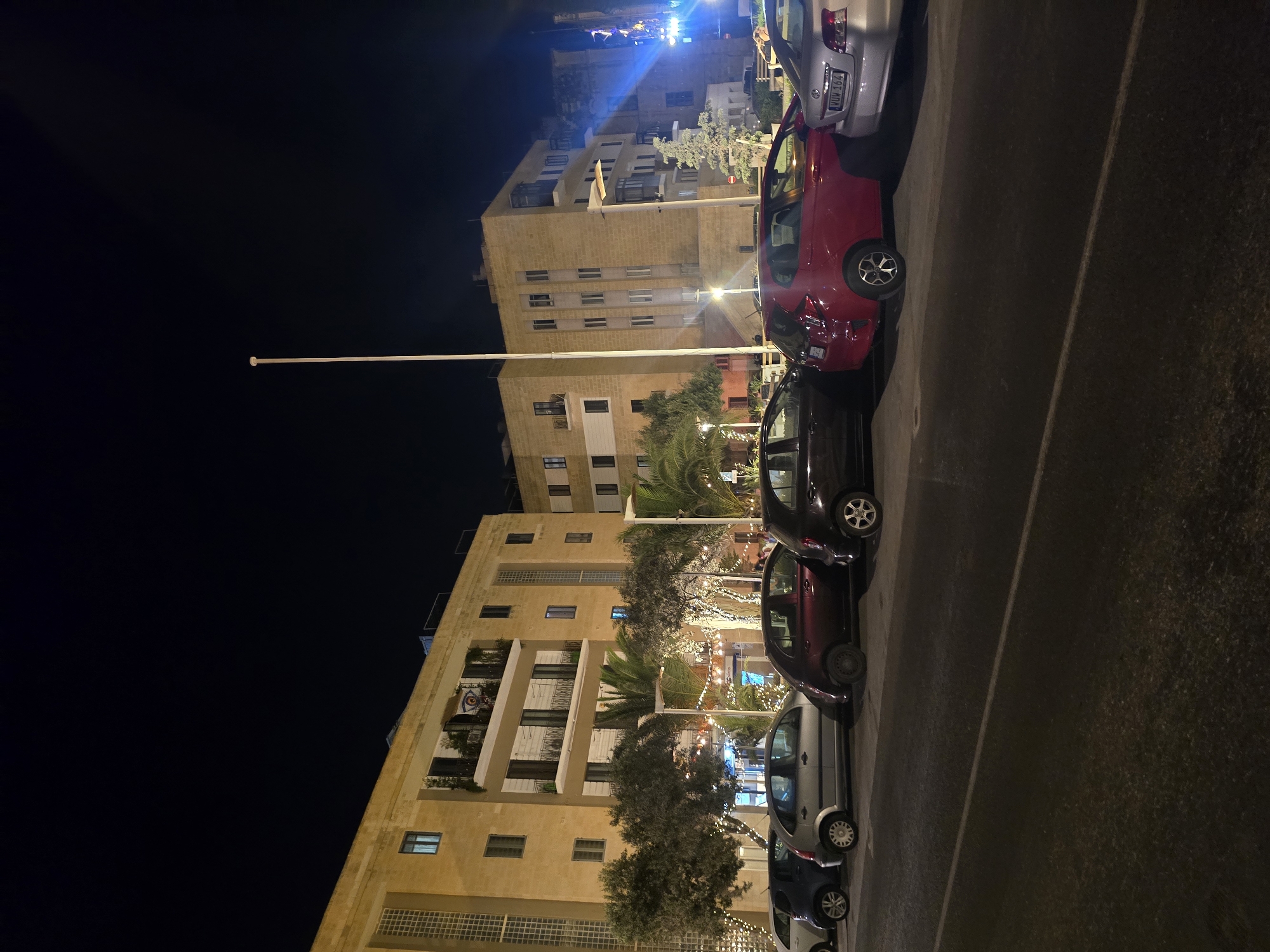 The image depicts a nighttime street scene in Valletta, Malta. Several cars are parked along the side of a road in front of a multi-story building. The building is illuminated by streetlights and lights from within the structure, revealing its beige facade and balconies. Decorative string lights are visible in front of the building, draped over trees and creating an festive atmosphere. The sky is dark, indicating it is nighttime.