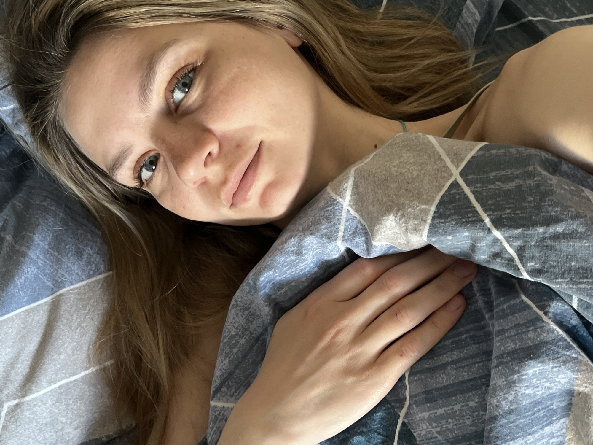 A woman with fair skin and light brown hair lies in bed, partially covered by a blue and grey patterned comforter. Her face is turned slightly to the left, with her eyes gazing upward. Her right hand rests on the comforter near her chest. The scene is indoors, likely a bedroom. There's no specific information to indicate this image was taken in the city of Chaikovsky, Russia.