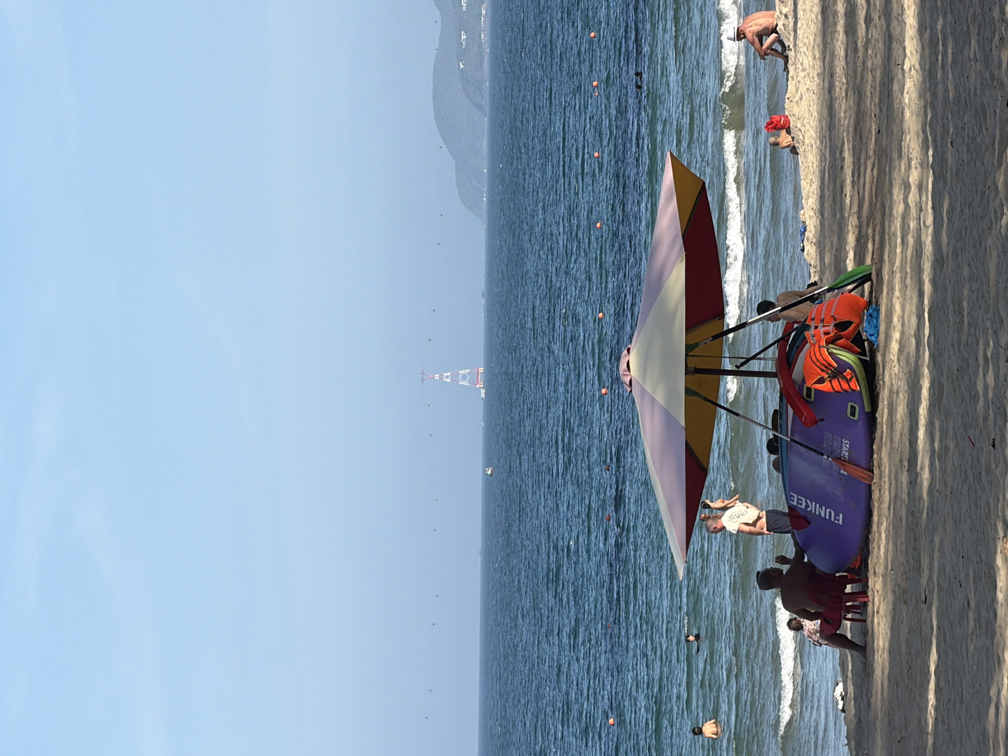This image captures a section of a sandy beach in Nha Trang, Vietnam, with the ocean and sky dominating the background. In the foreground, a large, colorful umbrella provides shade for several people and beach equipment. A purple stand-up paddleboard labeled "FUNKEE" is visible, along with orange life vests and paddles.
Several individuals are present on the beach. Some are seated under the umbrella, while others are standing or walking. In the water, a few people are swimming or wading. The ocean is a deep blue, with moderate waves breaking near the shore.
In the distance, a tall, red and white structure, possibly a tower or pylon, stands in the sea. Hills and a hazy coastline are visible on the horizon. The sky is a clear, pale blue. The overall scene depicts a relaxed day at the beach with typical recreational activities.