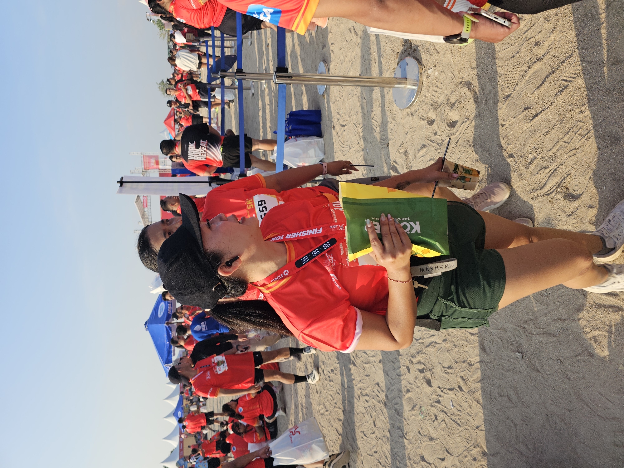 The image depicts a sunny outdoor scene, likely a race event, taking place on a sandy surface with tire tracks and footprints visible. In the foreground, a woman wearing a black baseball cap and a red t-shirt with "CIRCLE K" branding is holding a green and yellow bag and a plastic cup with a straw. She has earbuds in her ears and is wearing dark green shorts and white athletic shoes. Behind her, other individuals in red shirts are present, some with bib numbers. In the background, tents, barriers, and more participants can be seen under a clear blue sky. The presence of "CIRCLE K" branding suggests a possible sponsorship or partnership for the event. Based on the provided crops which show tents with "AQUA" branding, a common water brand in Indonesia, and the sandy, beach-like terrain often found along the coast, this event could be located in Denpasar, Indonesia, likely near a coastal area like Sanur or Kuta which are popular for such activities.