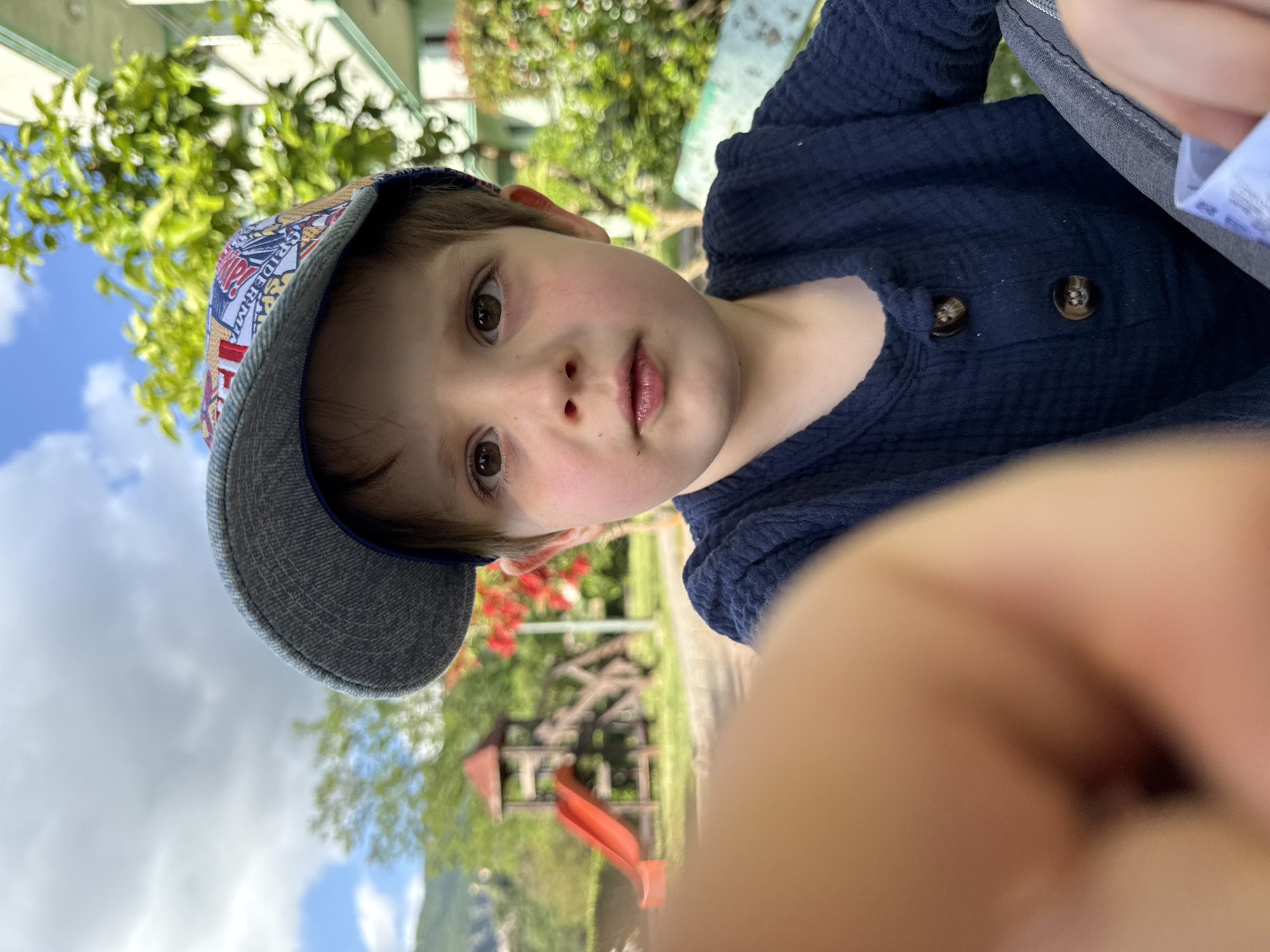 A young Caucasian boy with light brown hair and brown eyes wears a baseball cap with a denim brim and a cartoon print on the crown. He is also wearing a navy blue button-down shirt. The boy is looking directly at the camera. The image appears to have been taken from a low angle, possibly by the boy himself.
In the blurred background, a playground with a red slide can be seen. There is also lush greenery, including trees and flowers. The photo may be taken in Zelenika, Montenegro, considering the prompt's request. The sky with scattered clouds can be seen above the trees.