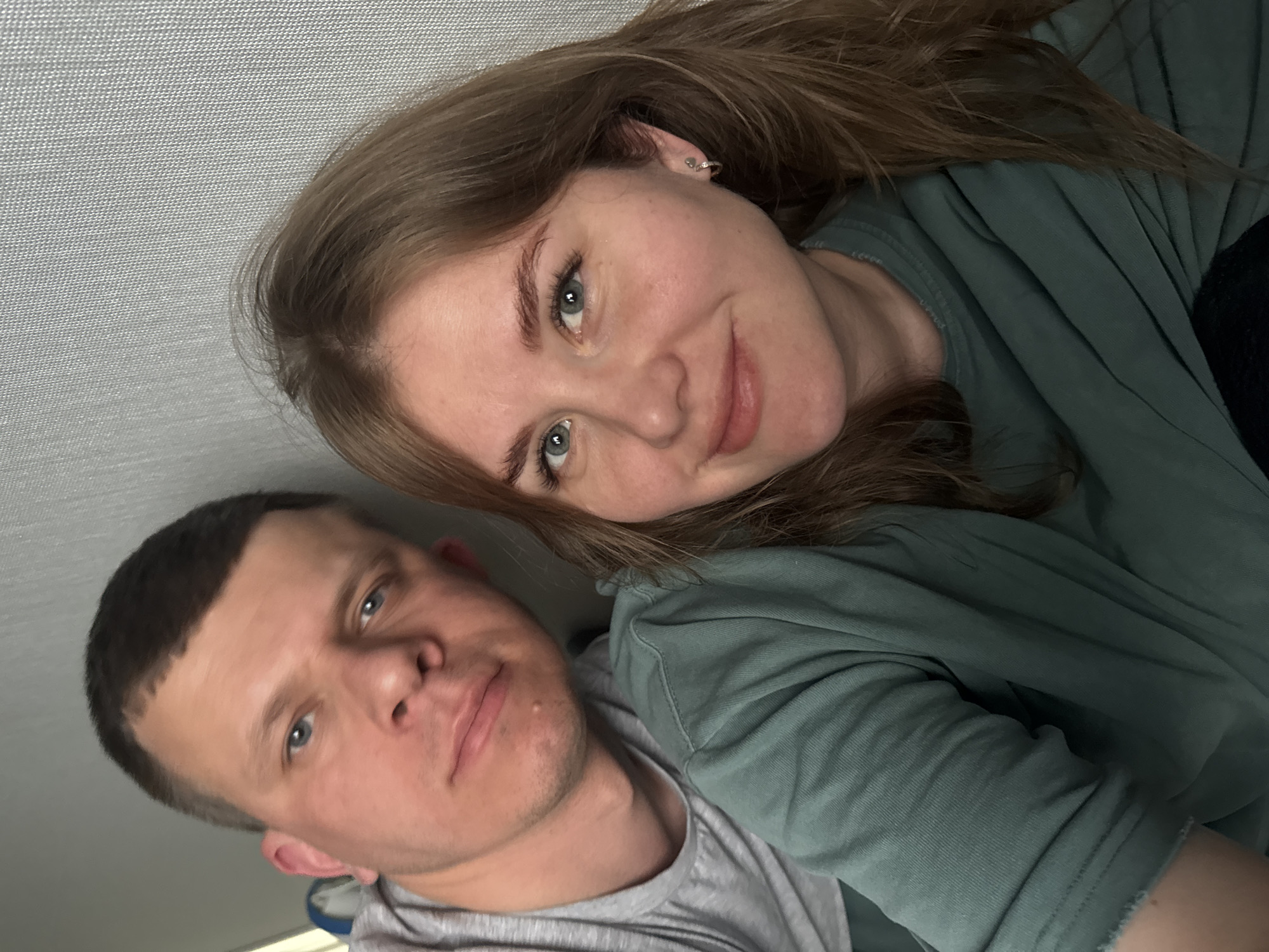 Two individuals are depicted in an indoor selfie. A woman with long brown hair and fair skin is positioned on the right side of the frame, wearing a green top. She is slightly smiling. To her left is a man with short, dark hair and fair skin, wearing a grey t-shirt. The background appears to be a plain, light-colored wall. The photo appears to be taken at an angle. There is no information to determine where the photo was taken in the city of Ферма, Russia.