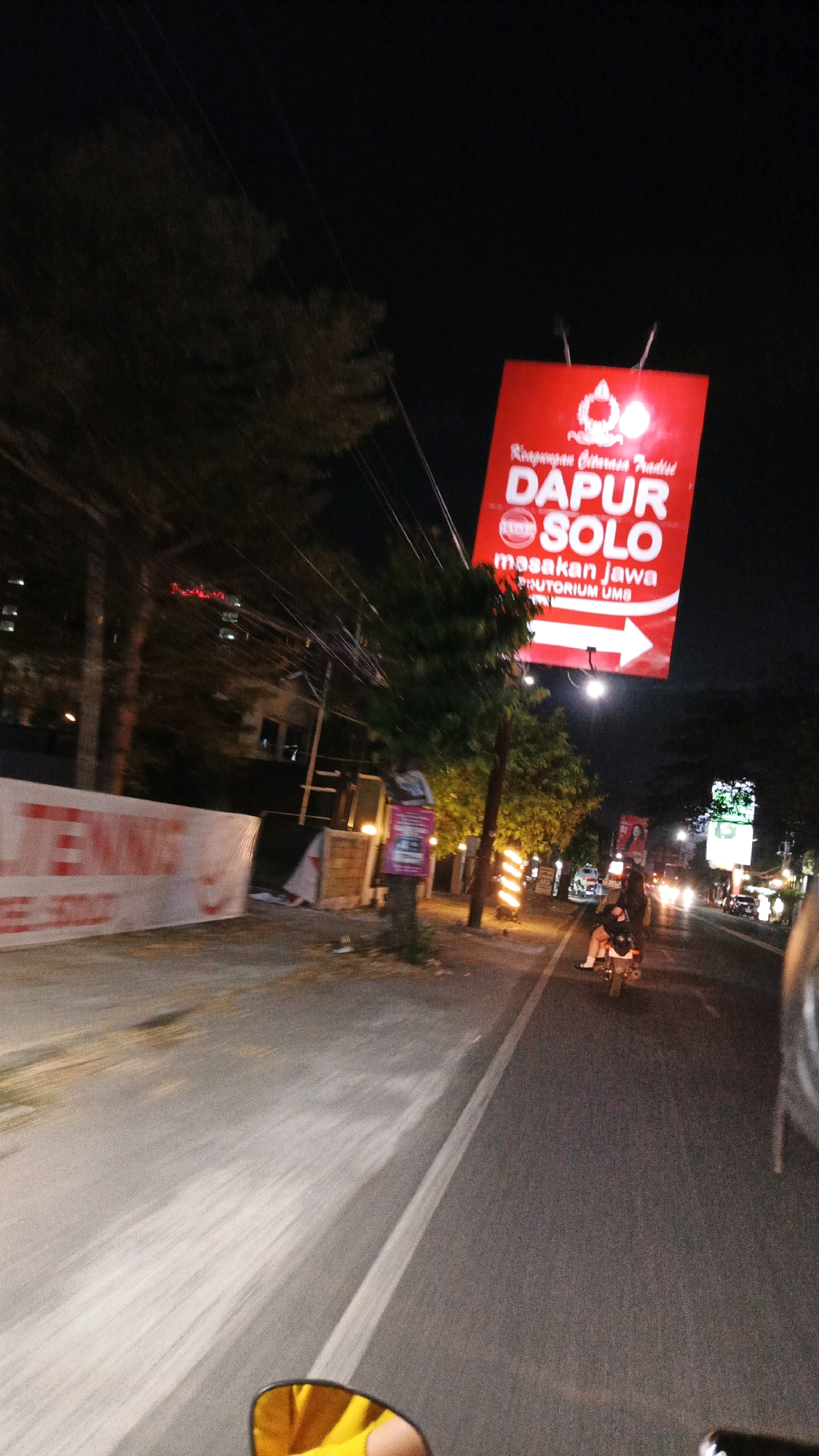 The image captures a night scene on a street in Blulukan, Indonesia, likely from the perspective of someone riding a motorcycle. Dominating the upper right of the frame is a large, red, illuminated sign that reads "DAPUR SOLO" in large white letters, with smaller text above and below indicating "Riesmpanan Citarasa Tradisi" and "masakan Jawa" respectively. Below "masakan Jawa" is smaller text "FRUTORIUM UMS" and a white arrow pointing to the right.
The street is paved with asphalt and marked with a white line. To the left of the road, a blurred banner with red lettering that is partially legible as "LENING" is visible. Further down the street, trees with illuminated foliage and streetlights create a warm glow. Another smaller, illuminated sign with green text is visible further in the distance.
A person is riding a motorcycle in the middle of the right lane, illuminated by streetlights. Their legs and the back of the motorcycle are visible. The foreground shows a blurred yellow reflection in what appears to be a motorcycle's rearview mirror, suggesting the photographer is also on a motorcycle. The overall atmosphere is that of a bustling urban night, with traffic and illuminated signage creating a dynamic scene.