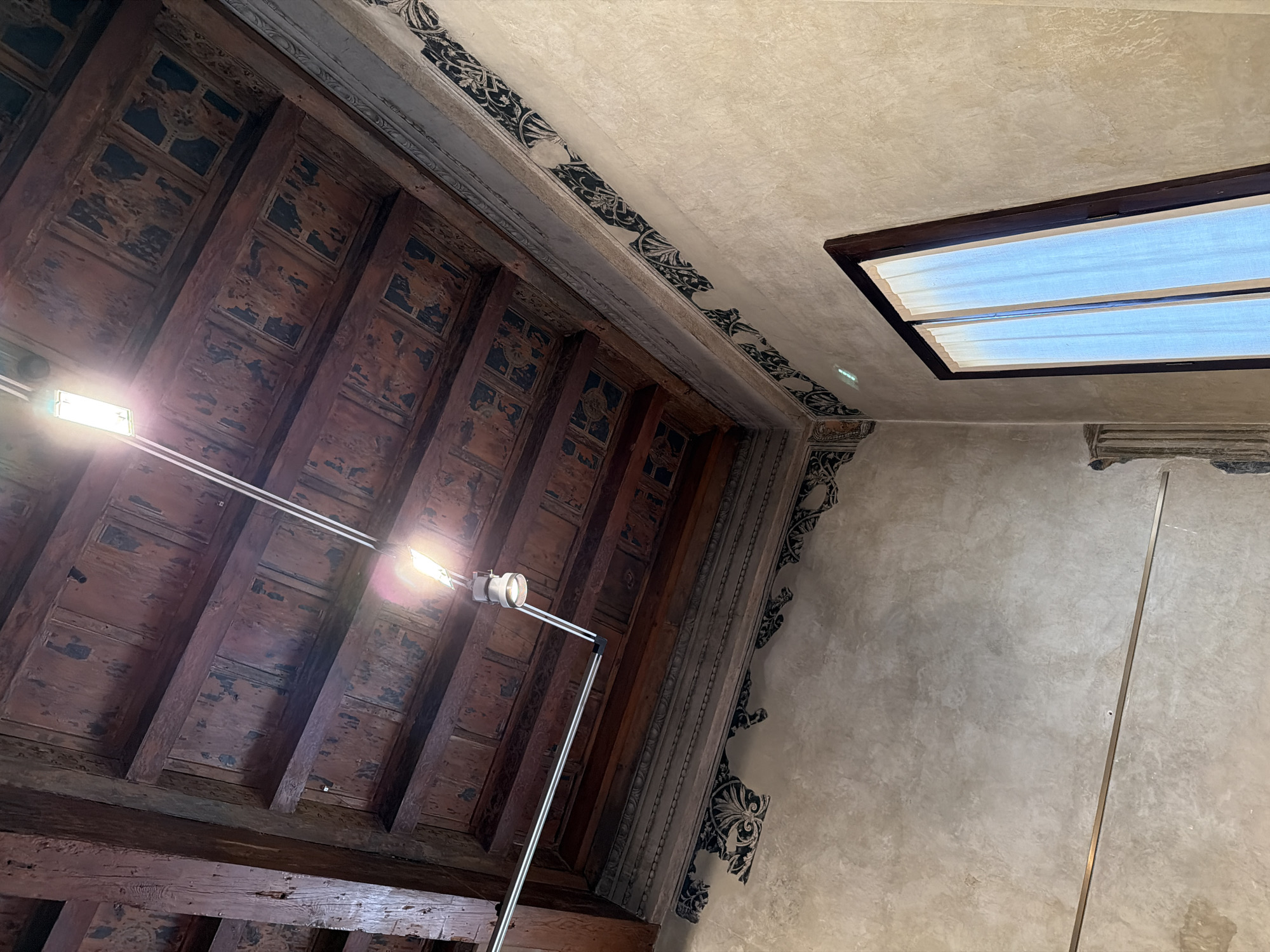 The image shows a view looking upwards inside a building in Mantua, Italy. The left portion of the frame is dominated by a dark, ornately decorated wooden ceiling with a repeating pattern of squares and beams. A modern, adjustable floor lamp with two spotlights stands in front of the ceiling. To the right of the ceiling is a plain, textured wall. The top right corner contains a rectangular skylight with a bright, translucent panel. The wall features decorative, ornate trim at the junction with the ceiling. The overall scene is a combination of historical architectural elements and modern lighting.