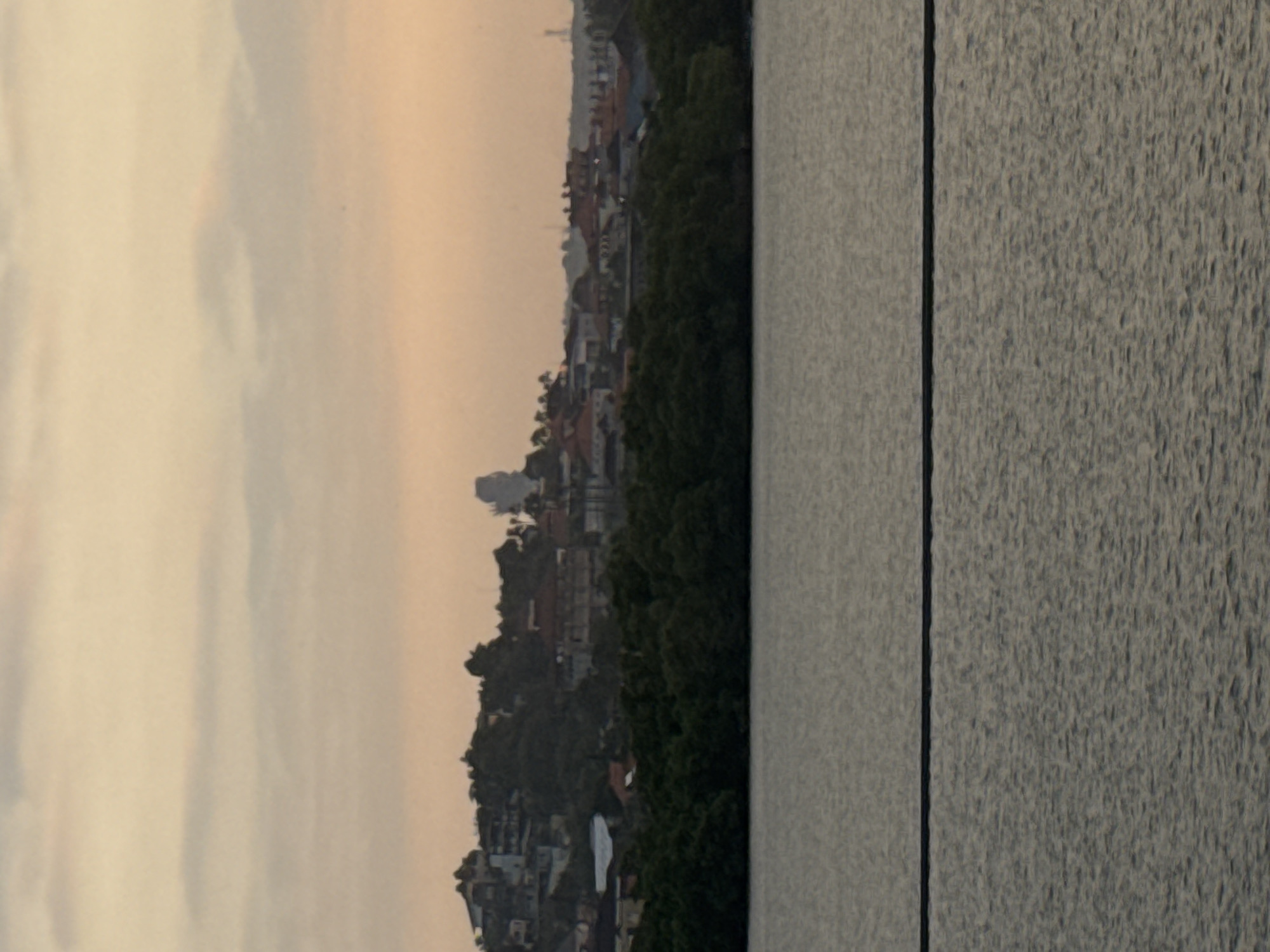 The image depicts a tranquil, hazy sunset over the water, with a densely vegetated coastline in the middle ground and a cityscape on the hills beyond. The water in the foreground is calm and reflects the soft hues of the sky, which transitions from pale yellow at the horizon to a muted orange and pink overhead. A dark, horizontal line, likely a cable or wire, stretches across the water's surface in the lower portion of the frame.
The middle ground is dominated by a thick band of dark green foliage, suggesting a mangrove forest or dense coastal vegetation. Behind this, the hillside is covered with a mix of buildings and trees. The buildings are varied in style and color, with red-tiled roofs being prominent. The cityscape appears somewhat scattered, with structures built into the sloping terrain. A distinct, large, light-colored, silhouette of a statue or monument is visible on the highest point of the hill in the left half of the image, partially obscured by trees. Further to the right, a few more buildings and what appear to be communication towers or antennas are discernible on the distant horizon. The overall atmosphere is serene and somewhat diffused due to the sunset light and potential atmospheric haze. This scene is consistent with a view of the coastline near Kuta Selatan, Bali, Indonesia, known for its blend of natural beauty and developing urban areas.