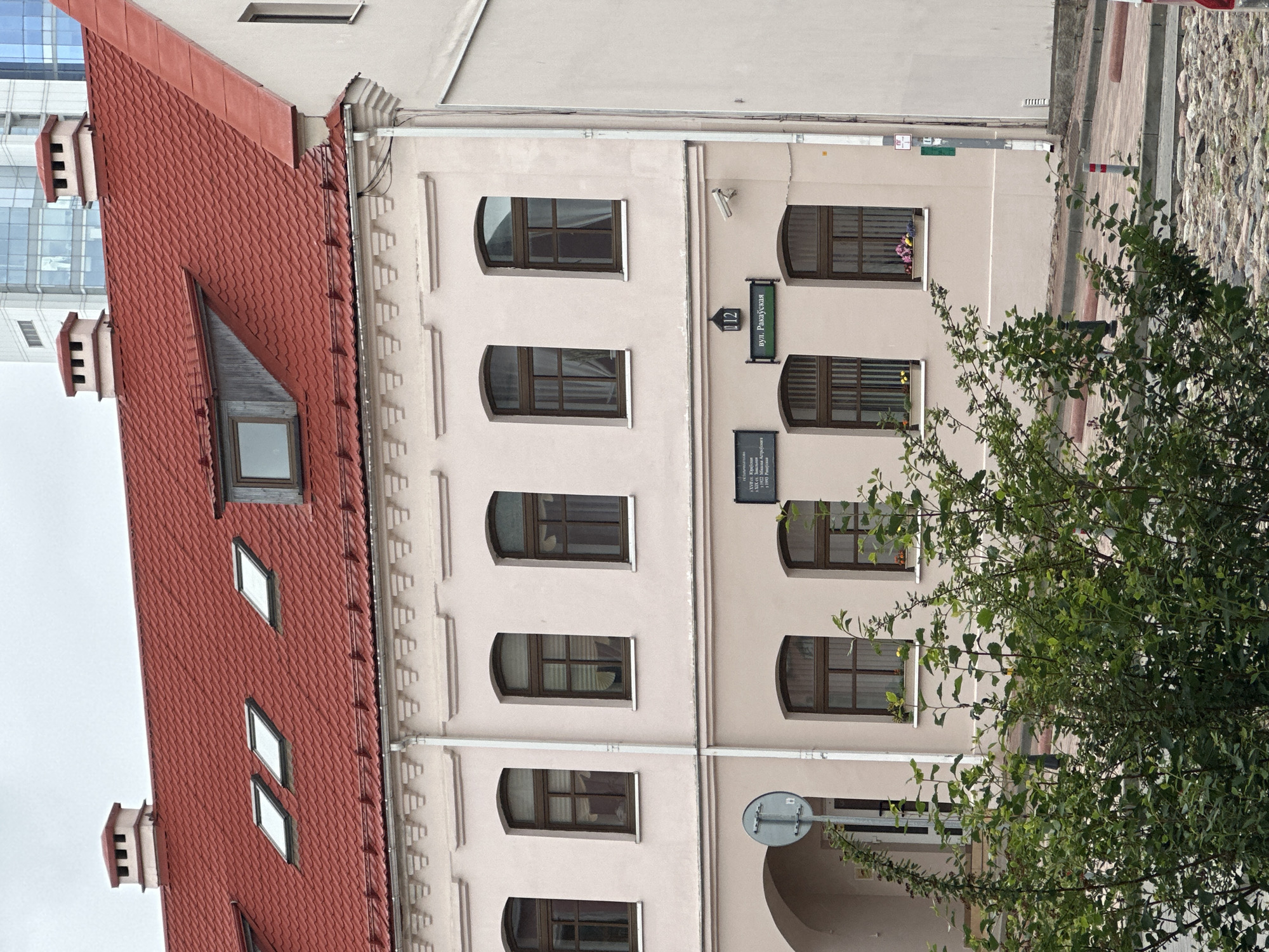 The image shows the facade of a light-colored building with a red tiled roof. The building has several arched windows with brown frames on the lower two floors. There are three skylights on the roof, and two chimneys. A street sign indicates "вул. Ракаўская" and the building number "12". There is also a sign with Cyrillic text above the street sign. A security camera is mounted on the wall. In the foreground, there is a dense bush with green leaves. The street in front of the building is paved with cobblestones. The overall scene suggests an urban environment in Minsk, Belarus.