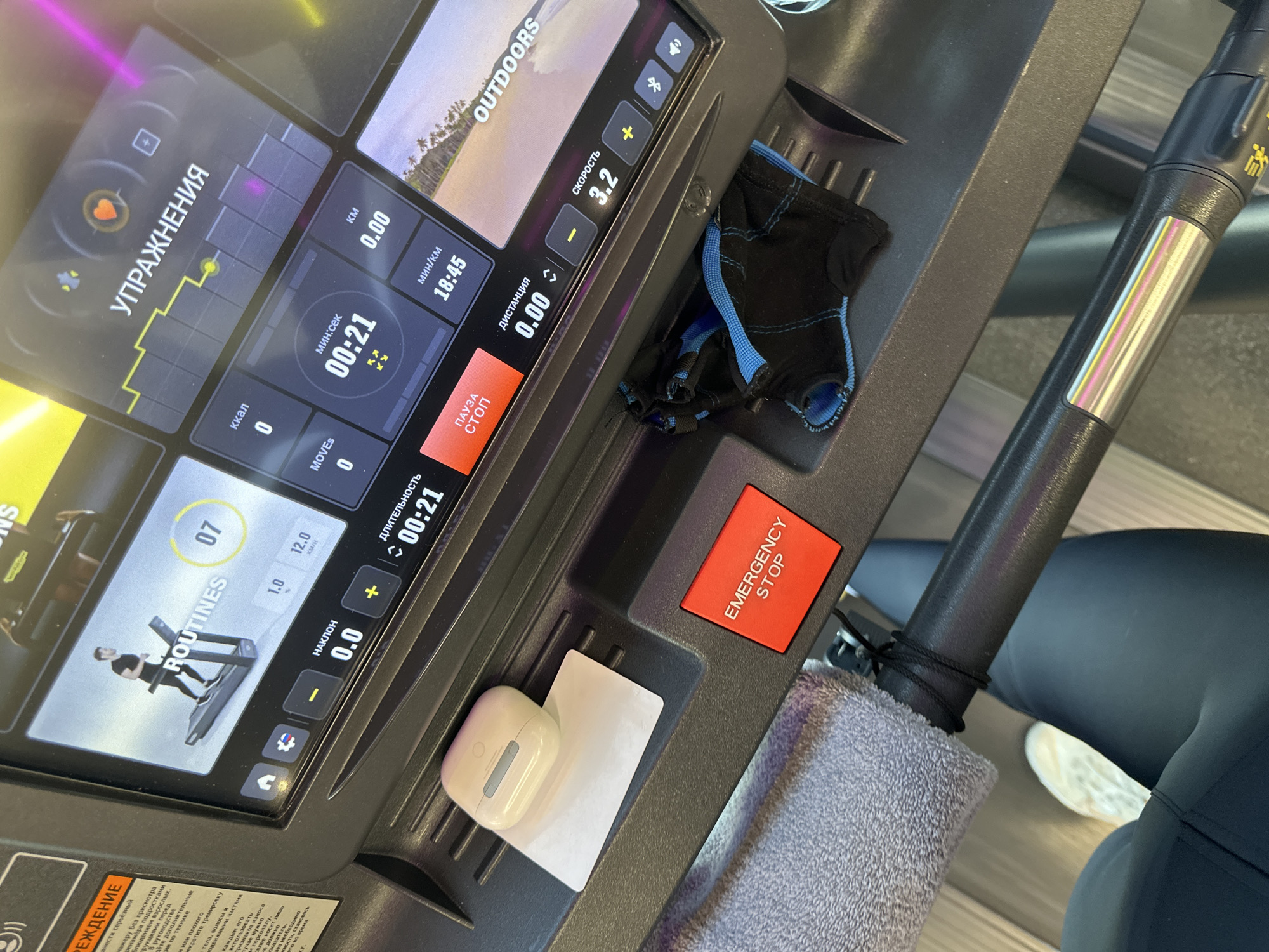 The image shows the console of a treadmill, likely inside a gym or fitness facility, though the exact location in Perm, Russia, is not discernible from the visual information alone. The treadmill display shows that the workout is ongoing, indicating a time of 00:21 (minutes and seconds). The speed is set at 3.2, and the distance covered is 0.00. The incline is at 0.0. There is a routine program displayed on the console. A pair of black and blue fingerless gloves rests on the console area. To the side, one can see a pair of wireless earphones and a white rectangular card. An "EMERGENCY STOP" button is visible. A gray towel is draped over the handle of the treadmill. The lower portion of an individual wearing dark leggings and white sneakers is partially visible to the side of the treadmill handle.