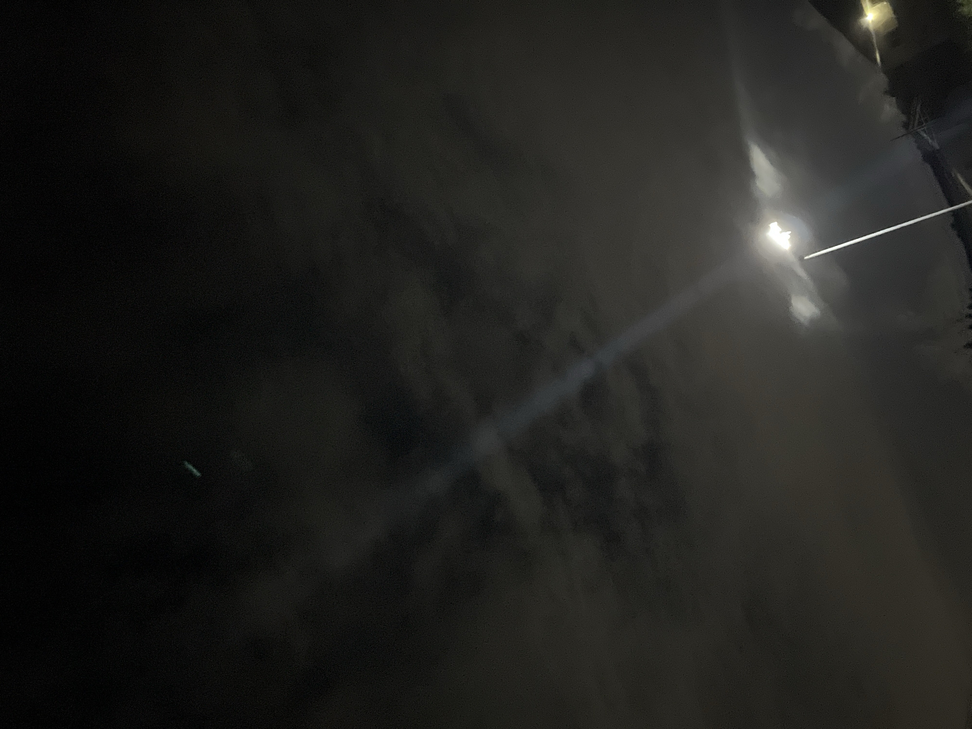 The image shows a night scene in what appears to be an urban area. A bright light source, possibly a street lamp, illuminates the area. There is a structure, possibly a building or a pole, visible in the upper right corner. The rest of the scene is dark, with some cloud-like formations that obscure much of the details. A beam of light cuts through the darkness, originating from the bright light source.