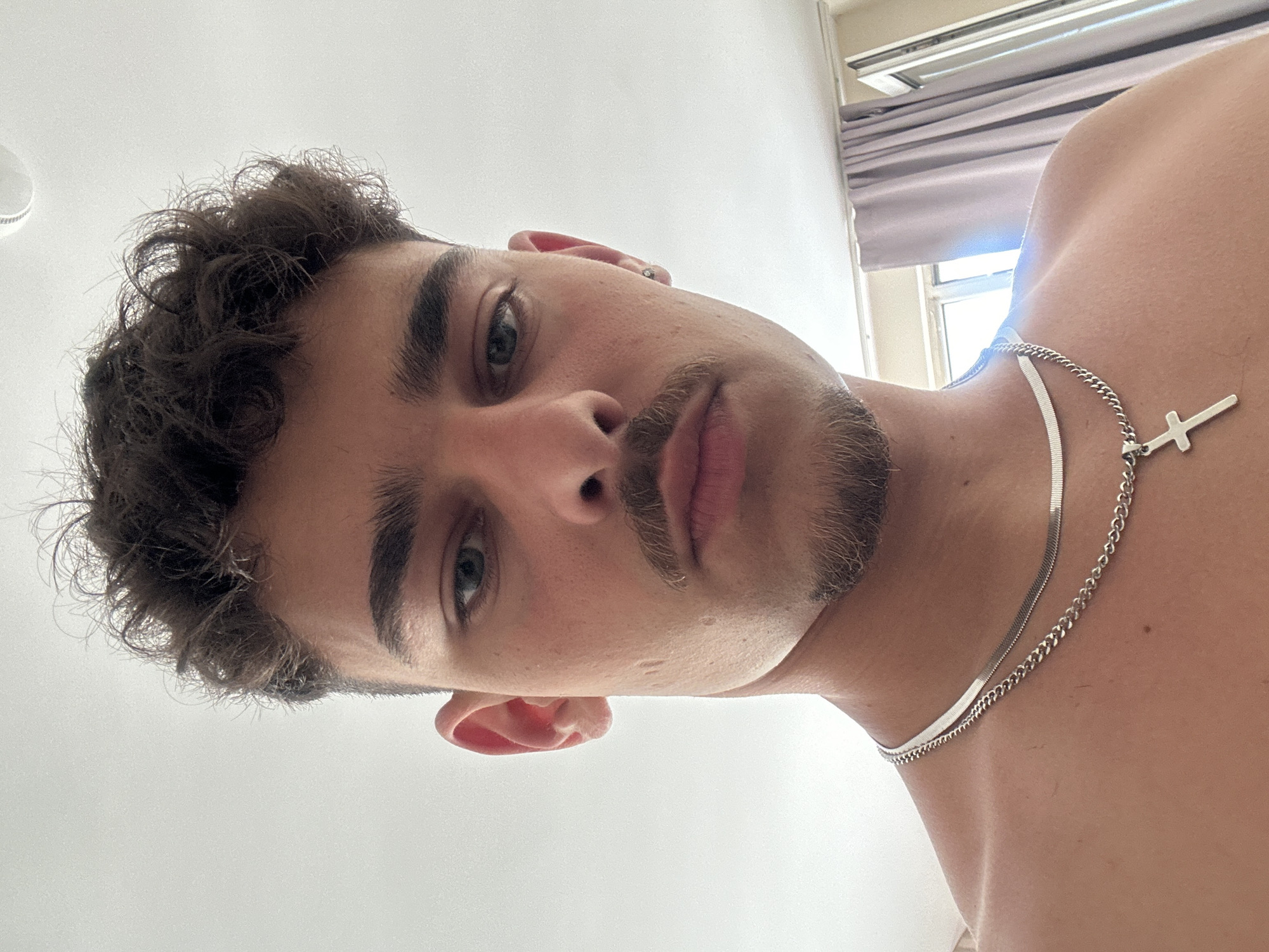 A medium-shot, low-angle selfie depicts a light-skinned male with dark, curly hair and a mustache/goatee combination. His eyes are green, and he has a piercing in his left ear. He is wearing two silver necklaces, one a herringbone style and the other a chain-link style with a cross pendant. He is shirtless, and the background shows a ceiling, a window with a gray curtain, and a light source that appears to be daylight. There is no information to suggest the location is Sofia, Bulgaria.