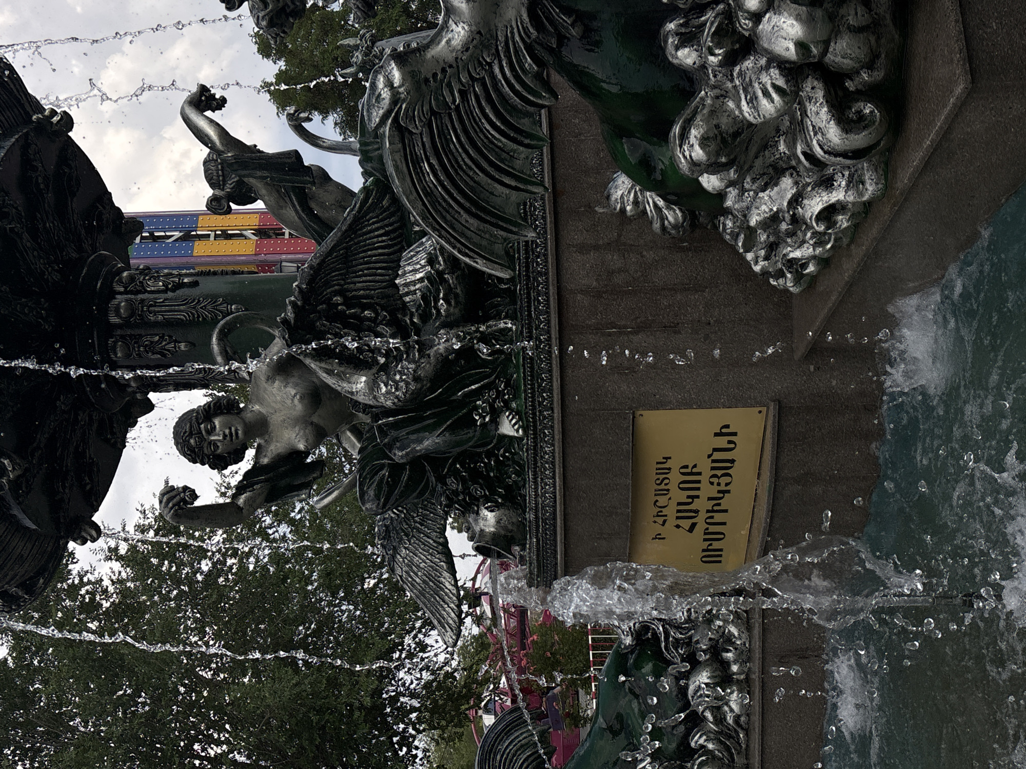 The image shows a detailed sculpture of a fountain in Yerevan, Armenia. The sculpture features several winged figures and mythological creatures, intricately designed and appearing to be made of bronze or a similar dark metal. Water is actively flowing over the sculptures, creating a dynamic and glistening effect. In the background, elements of an amusement park are visible, including a colorful Ferris wheel or roller coaster structure, suggesting the fountain is located in a public entertainment area. A golden plaque with Armenian script is affixed to the base of the fountain. The overall scene captures a blend of artistry and leisure, set against a cloudy sky and lush green trees.