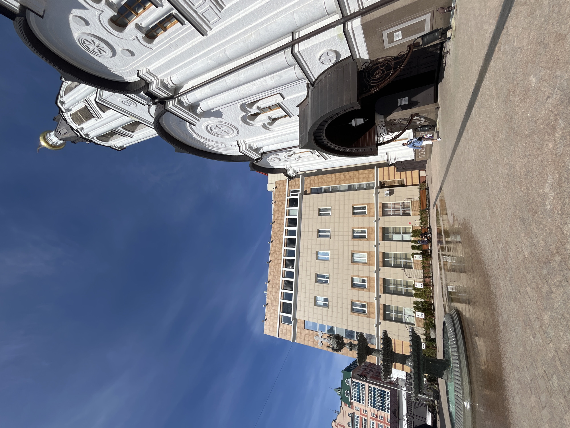 This image captures an outdoor urban scene in Ulyanovsk, Russia, under a bright, clear blue sky with a few wispy clouds. The setting appears to be a city square or pedestrian area, characterized by a wide expanse of light-colored, wet-looking paving in the foreground and midground, reflecting the sunlight.
On the left, a grand, white architectural structure, likely a church or cathedral, stands prominently. It boasts an ornate and classical design with multiple tiers, columns, arched windows, and circular decorative motifs. A golden dome topped with a cross rises majestically towards the sky, while another large dome with decorative elements is partially visible. Near the entrance of this building, a decorative black wrought iron gate or archway with intricate scrollwork and a lamppost is present. A single person, dressed in light clothing, is seen standing near this gate, facing away from the camera.
To the right, a modern, multi-story building contrasts with the classical architecture. It features a light brown or tan facade with a grid pattern of numerous rectangular windows. Along its base, several benches are visible, with a few indistinct figures seated, and small green plants or hedges add a touch of greenery. Further in the foreground, a dark, multi-tiered fountain, possibly made of metal, can be seen, holding water. In the far background, glimpses of other buildings with varying architectural styles, including one with a green roof, complete the cityscape.
The scene depicts a typical daytime in a city, with people engaged in routine activities like walking or sitting. The clear sky and distinct shadows indicate sunny weather. No discernible text is visible on any signs or buildings.
