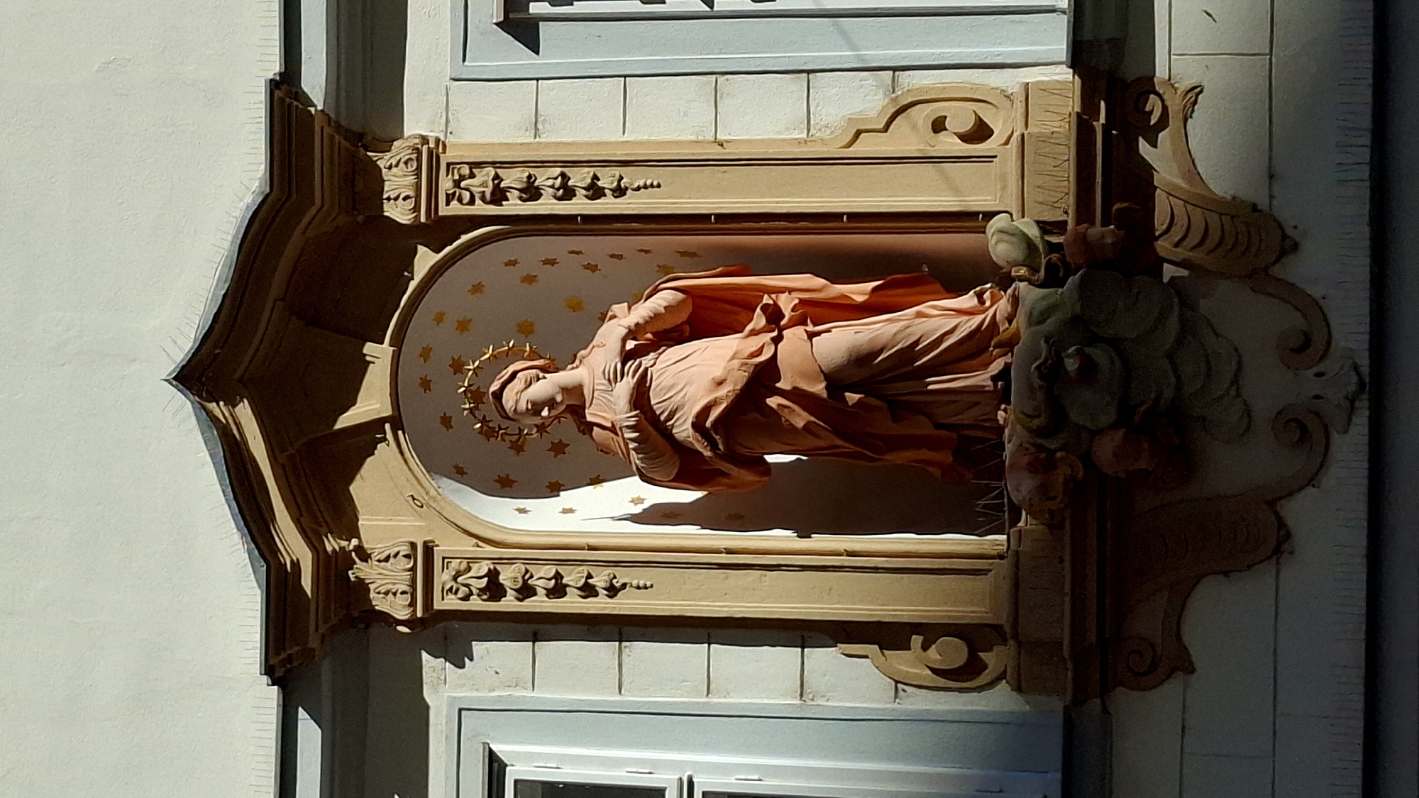 This is a photograph of a stone statue of the Virgin Mary located in an alcove on the facade of a building in Vienna, Austria. The statue depicts Mary with her hands clasped over her chest and a halo of stars above her head. She is wearing a draped robe. The alcove is ornate, with decorative columns and a patterned background. Below the statue, there is a decorative element resembling a cartouche with cherubic figures. The building's exterior is light-colored, with visible architectural details such as windows. The lighting suggests it is daytime, with sunlight casting shadows on the statue and the building.