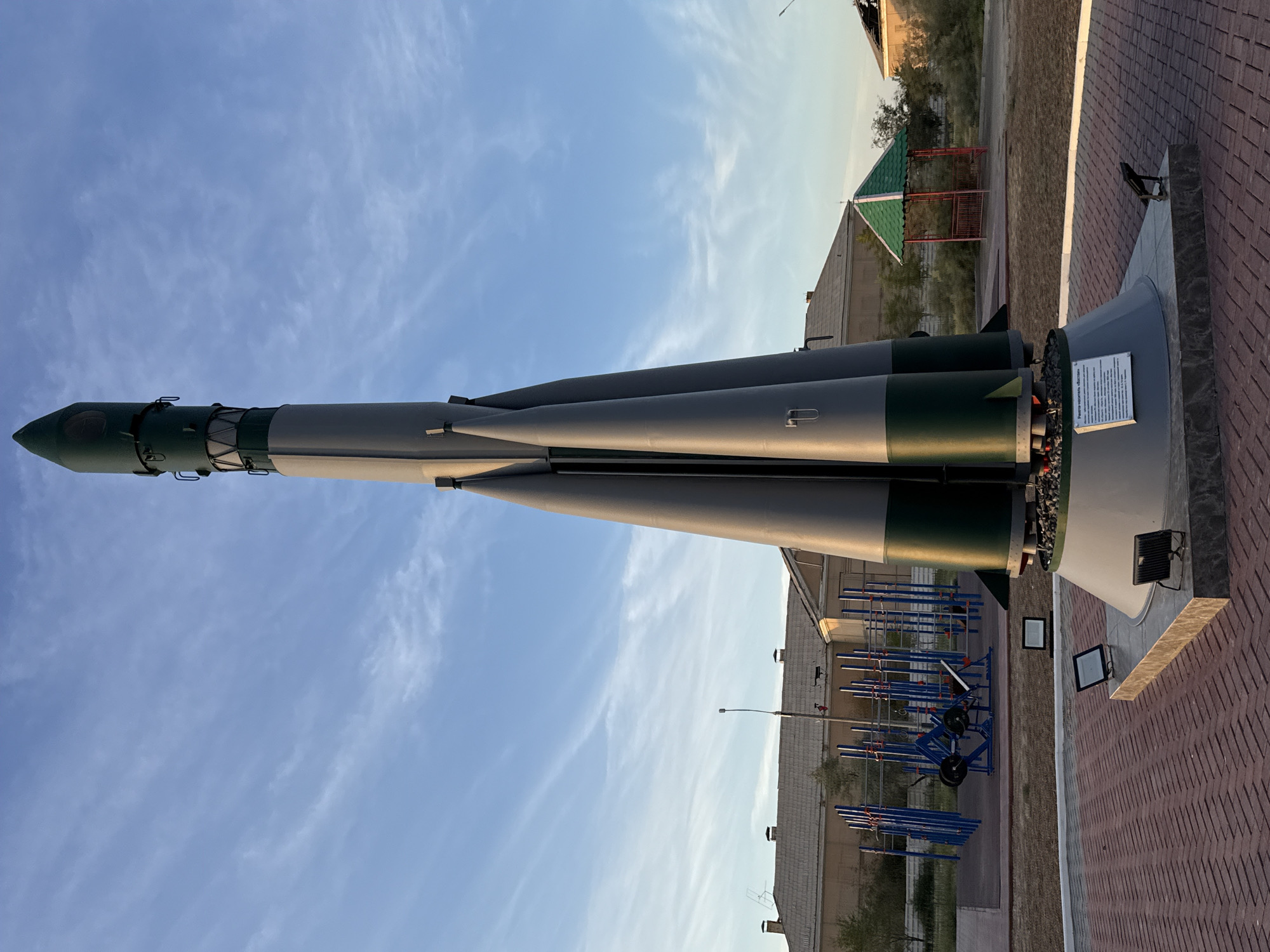 This is a full-frame, eye-level, outdoor photograph of a monument of the R-7 Semyorka rocket, a Soviet-era ICBM and space launch vehicle, displayed in the city of Baikonur, Kazakhstan. The rocket is depicted in its launch configuration, with four booster rockets surrounding a central core stage, all topped by a nose cone. The monument is painted in alternating sections of dark green and light gray. It stands on a circular, multi-tiered concrete base, which is surrounded by a paved area of red brick. Two small spotlights are positioned at the base of the monument on the brick pavement. A plaque with Russian text is affixed to the front of the monument's base.
In the background, there are several low-rise, beige-colored buildings with gray tiled roofs, typical of Soviet-era architecture. To the left of the rocket, there is an outdoor exercise area with blue and red metal equipment, including pull-up bars and weight machines. Further back, there is a small, green-roofed gazebo with red metal railings. The sky is a clear blue with thin, wispy white clouds. The lighting suggests it is late afternoon or early evening, with the sun casting long shadows. The environment appears to be a public park or square. There are no people or animals visible in the image.
