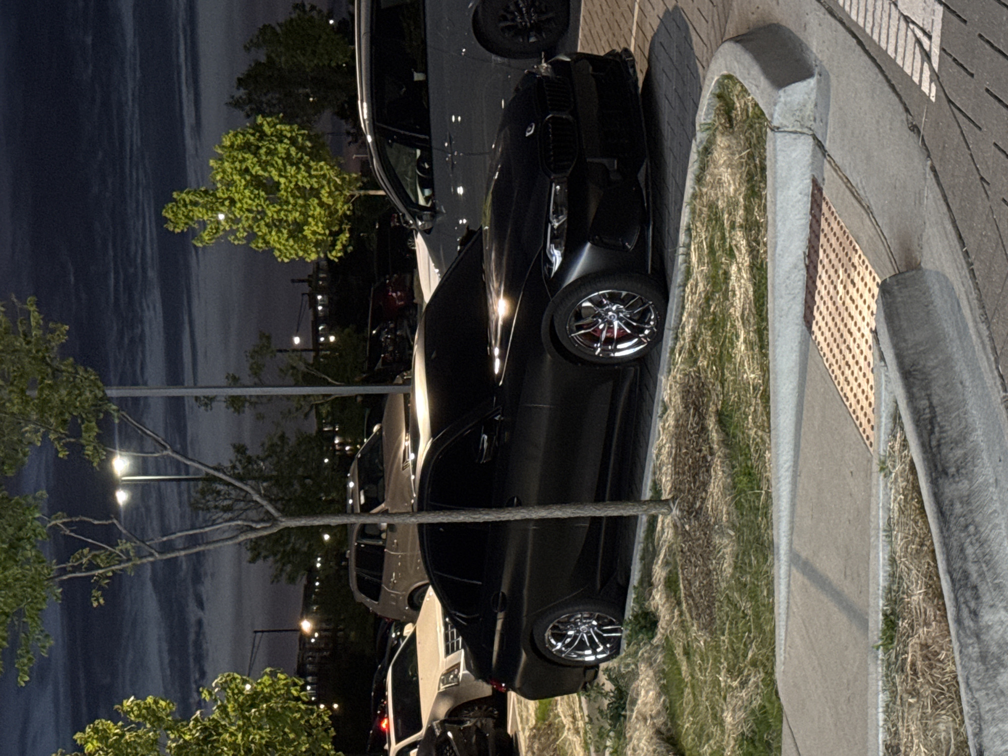 The photograph, taken at night in Schaumburg, United States of America, depicts a black car parked close to a sidewalk. The car's front driver's side tire is visible, showing a chrome rim. The sidewalk is made of concrete, with grass growing in the adjacent area. The sky is visible in the background, with streetlights illuminating the scene. A tree is on the left side of the image. Several parked cars are visible further in the background.