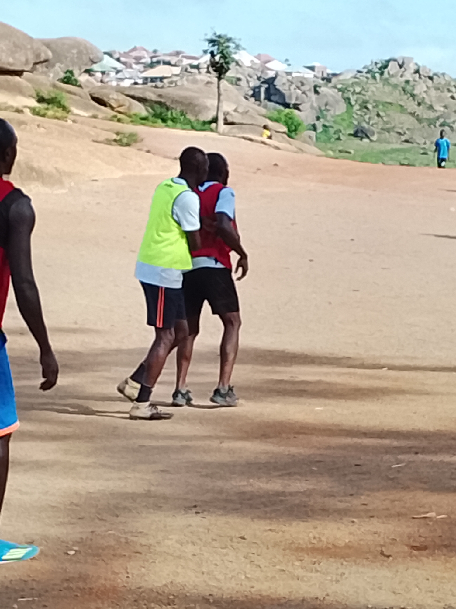 The image depicts a group of young men in an outdoor, dusty, and rocky environment, likely in Jos, Nigeria. In the foreground, the legs of three individuals are visible, all wearing athletic shorts and shoes. Two of these individuals are wearing vests, one bright green and the other red, suggesting they are engaged in some form of sporting activity or training. They are positioned close together, their bodies angled as if in motion or interaction. In the background, a hazy vista of a town is nestled amongst rolling hills and large rock formations characteristic of the Jos Plateau. A few buildings with distinctive rooflines are discernible. A single person in a blue shirt is visible standing further in the distance, and another small figure in a yellow top is seated on a rock. The overall impression is of a casual, perhaps recreational, gathering in a rugged, semi-urban landscape.