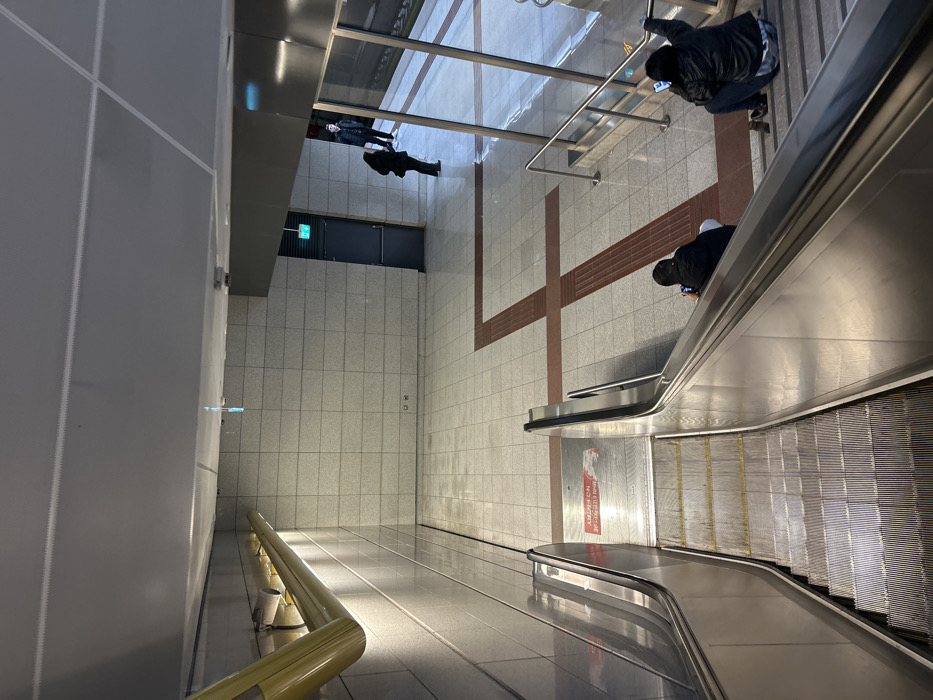 The image displays a spacious, modern interior, likely a metro station or a large public building in Nikea, Greece, viewed from an elevated perspective looking down towards a lower level.
Several individuals are present in the scene. On the upper level, near a glass barrier overlooking the escalators, one person in a dark jacket is seen, seemingly engrossed in a mobile phone. Below them, another individual wearing a dark jacket and a light-colored beanie or hood is seated or crouching on an escalator, also looking at a phone. Further back on the main concourse, two other people stand near a glass partition; one is in a dark coat, and the other is further away.
The setting features clean, minimalist architecture with light-colored tiled floors and walls. A prominent set of escalators descends from the mid-ground towards the bottom of the frame, characterized by sleek silver metal railings and a textured moving belt. A long, reflective wall on the left side of the frame creates a sense of depth, with a golden-colored handrail running along a lower section. The upper level is secured by a glass barrier, offering a clear view of the levels below. The main floor area is marked with distinct reddish-brown linear patterns, possibly indicating pedestrian pathways or designated zones. An illuminated green "EXIT" sign is visible on a wall in the background, above a dark doorway.
The activity in the scene suggests people are either transiting through the area, waiting, or engaged with their mobile devices, typical for a public transport hub. The lighting is bright and artificial, characteristic of an indoor public space, casting reflections on the polished floors and metal surfaces. On the floor adjacent to the escalator, a red rectangular sign with white text clearly reads "NO ENTRY" and "ΕΙΣΟΔΟΣ ΑΠΑΓΟΡΕΥΕΤΑΙ", which is Greek for "Entrance Forbidden." The overall atmosphere appears quiet and orderly.