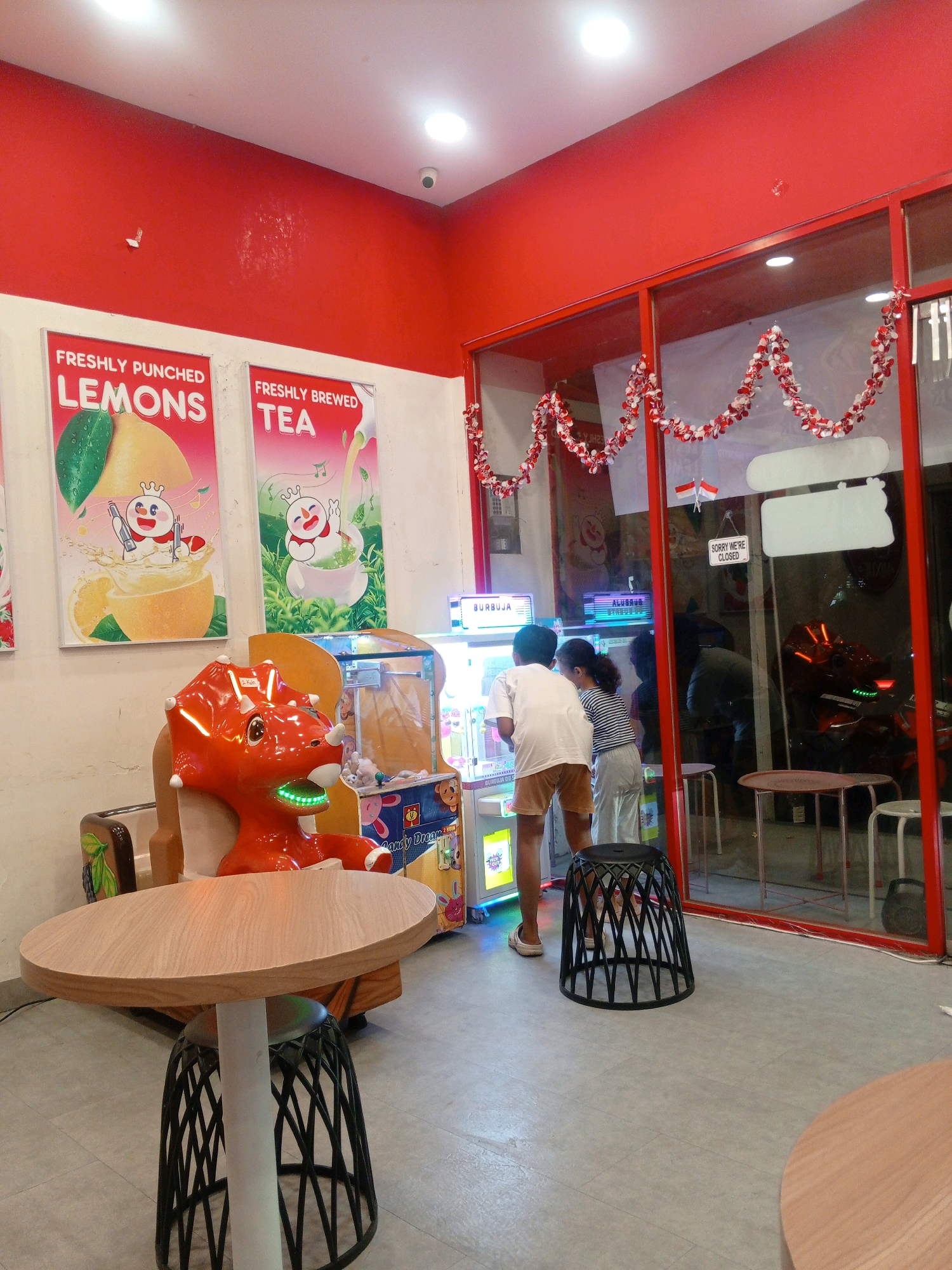 The image depicts the interior of a brightly lit establishment, likely a cafe or amusement center, with red walls and accents. On the left, two large posters advertise "FRESHLY PUNCHED LEMONS" and "FRESHLY BREWED TEA," featuring stylized cartoon characters. In the foreground, a red plastic dinosaur ride-on toy is positioned to the left of an arcade game. Two children, a boy in a white t-shirt and brown shorts, and a girl in a striped shirt and white pants, are interacting with the arcade game. The establishment has large glass windows with red frames, decorated with a string of red and white ornaments. Through the window, a motorcycle is visible. A round wooden table with a white pedestal base and a black basket-like stool sits in the foreground. The floor is tiled. A sign on the window reads "SORRY WE'RE CLOSED." The scene is in the city of Gabus, Indonesia.