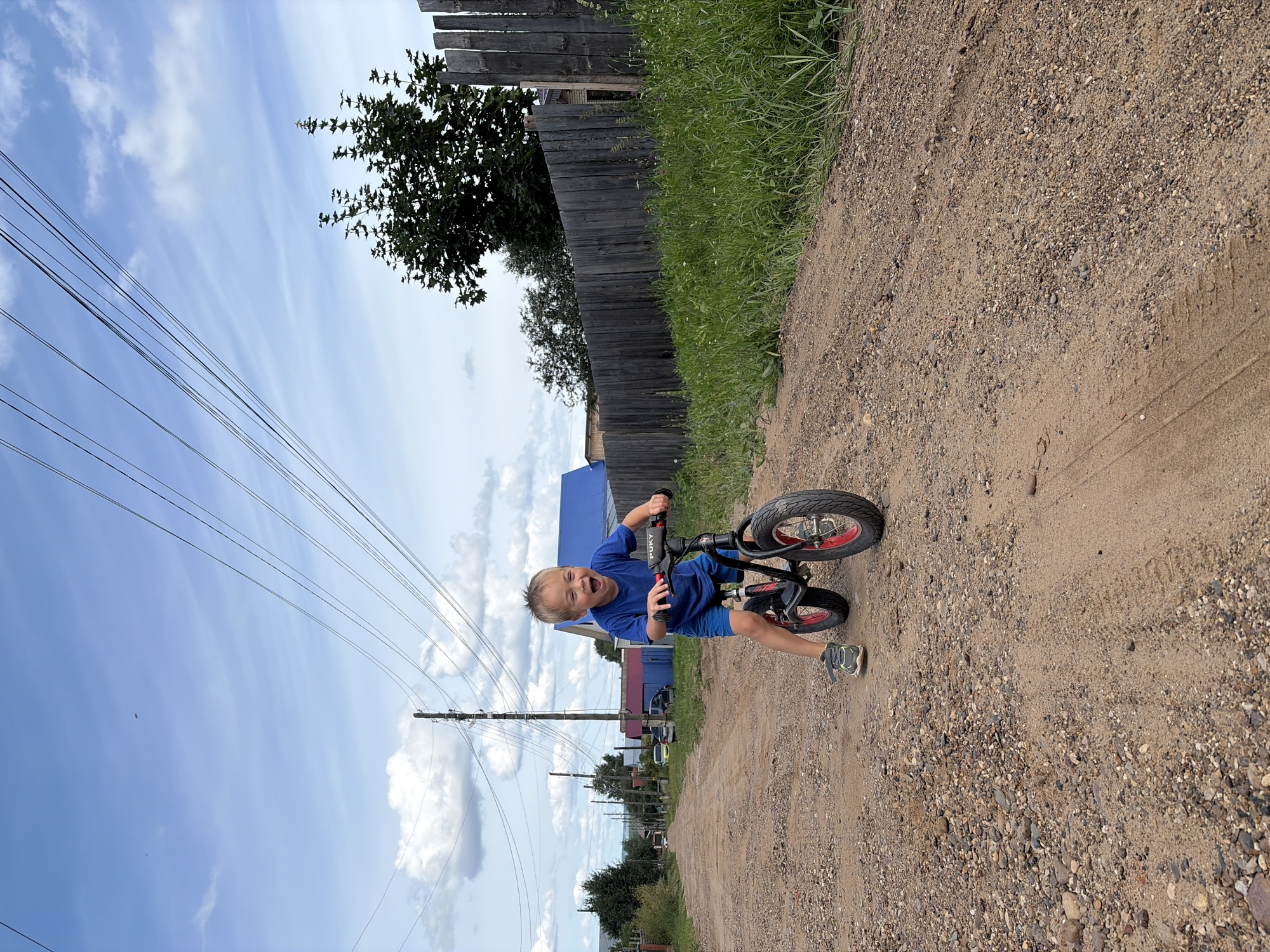 A young boy is riding a red and black balance bike on a dirt road in Olkhovka, Russia. The boy is wearing a blue t-shirt and blue shorts. He has blonde hair and is smiling with his mouth open. The road is lined with grass and a wooden fence on the right side. In the background, there are houses and power lines. The sky is blue with white clouds.